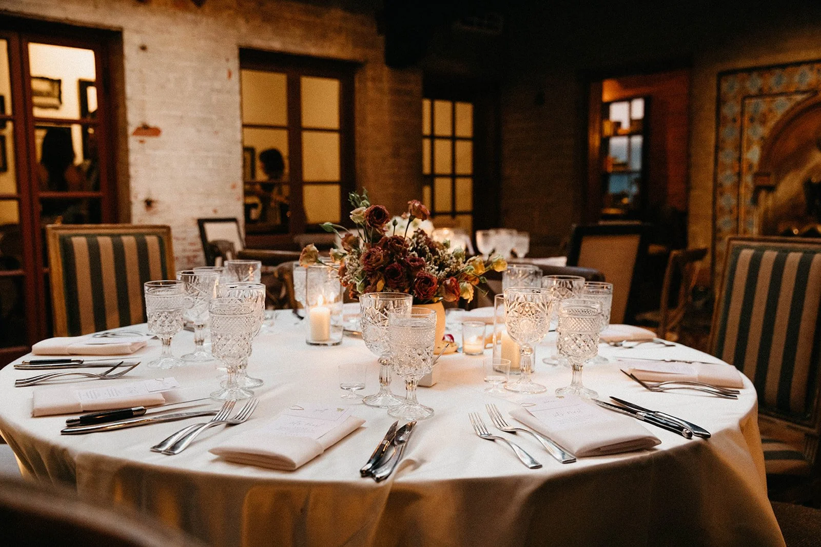 A banquet table set for a formal dinner with white tablecloth, crystal glasses, silverware, white napkins, and a central floral arrangement with candles in a warmly lit room.