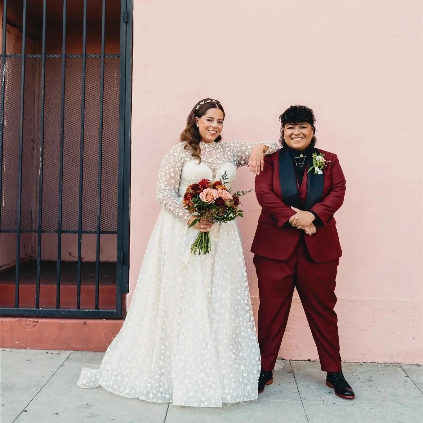 Two women standing and smiling in front of a pink wall. One is in a white wedding dress holding a bouquet, and the other is in a red suit.