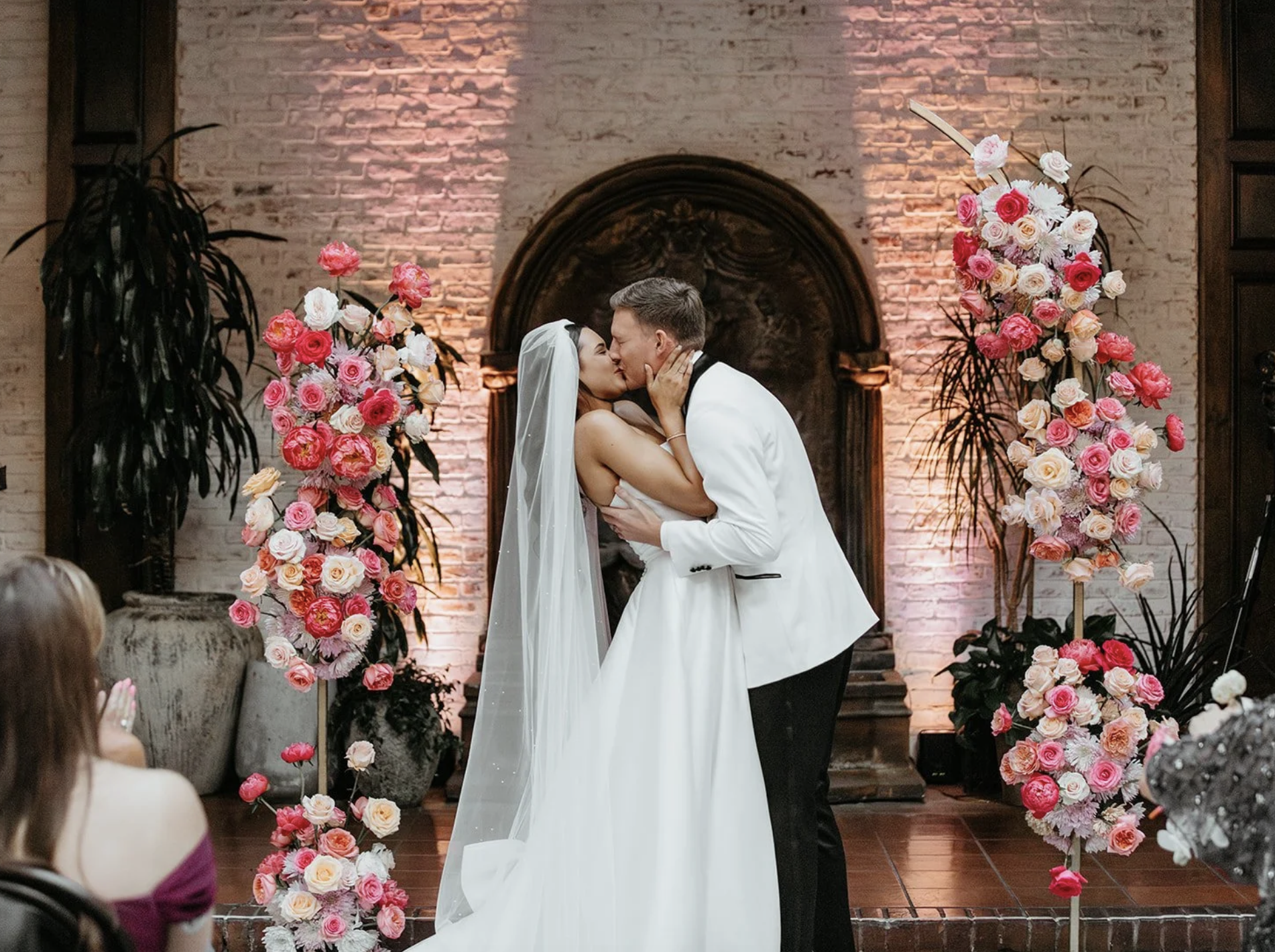 A bride and groom sharing a kiss during their wedding ceremony, surrounded by tall floral arrangements with pink and white roses, set against a brick wall with warm lighting.