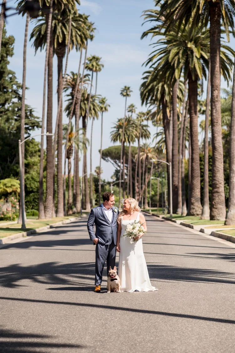 A newlywed couple in wedding attire, holding hands and smiling at each other on a tree-lined street with tall palm trees, accompanied by a French bulldog, under a bright sky in a sunny outdoor setting.