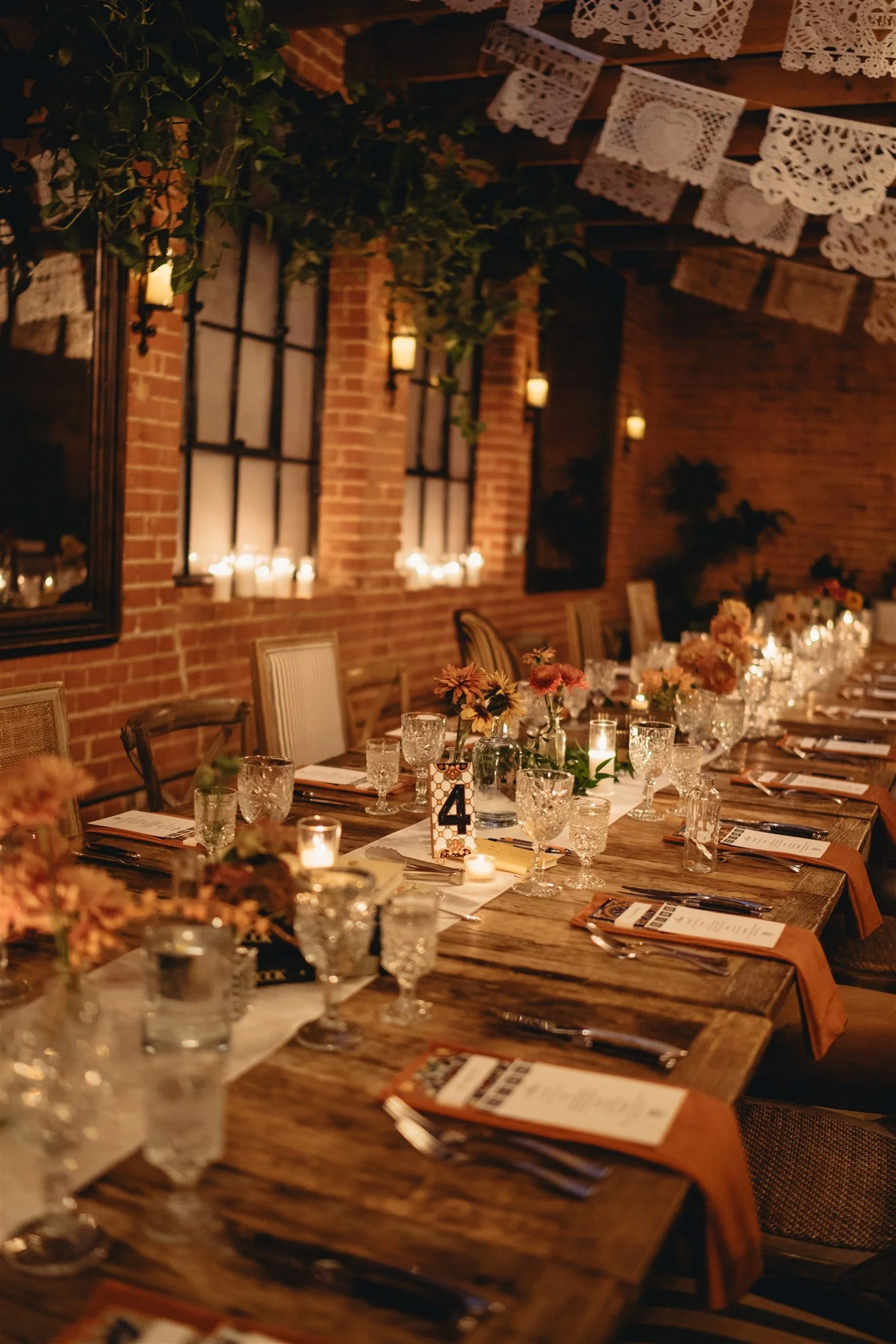 A cozy, rustic banquet hall decorated for a celebration with a long wooden table set with glassware, floral centerpieces, and candles. The room features exposed brick walls, large windows, and vintage-style hanging lace banners for decoration.