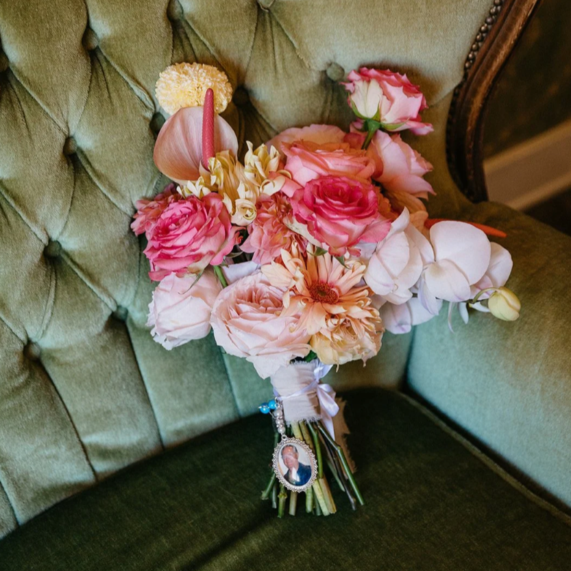 A bouquet of pink and white flowers with a small framed photo and beads hanging from the ribbon, placed on a green tufted armchair.