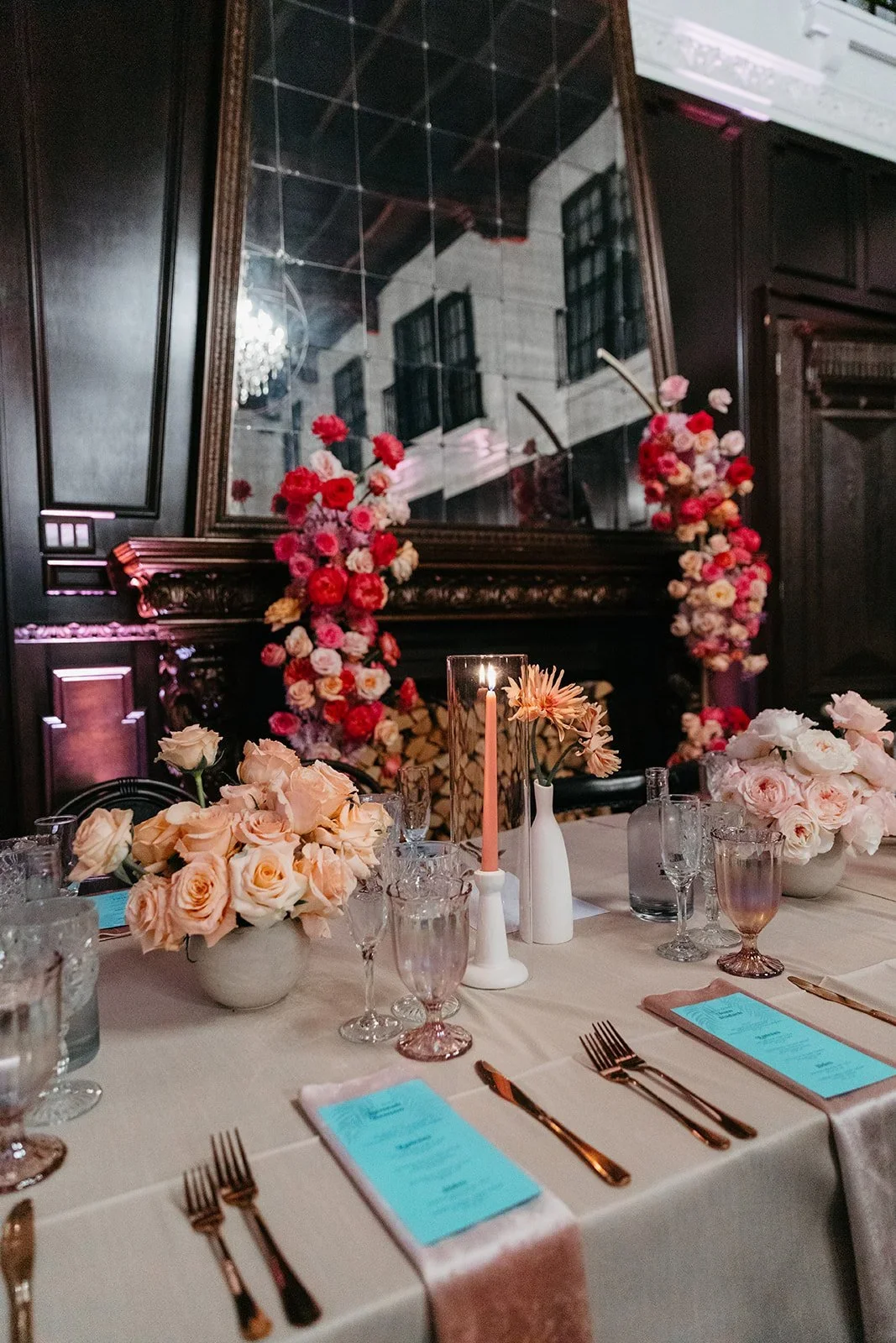 Elegant dining table set with pink and cream roses, floral centerpieces, candles, and place settings, in a room with dark wood paneling and floral arrangements behind a large mirror.