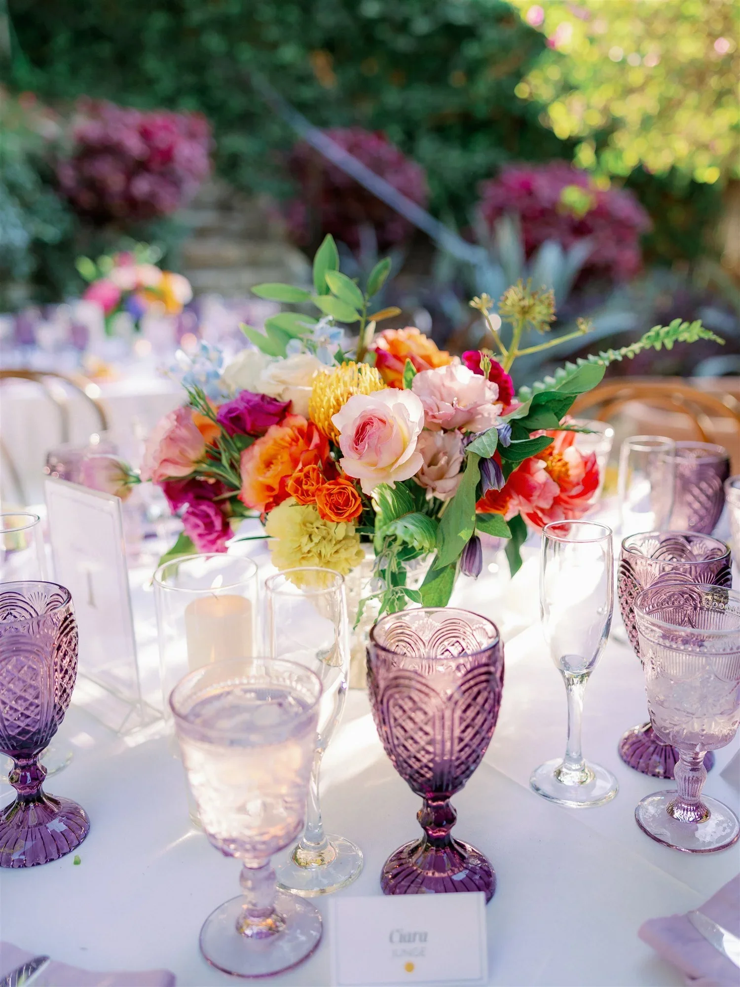 A table centerpiece with a large colorful floral arrangement of pink, yellow, orange, and purple flowers, surrounded by purple and clear patterned glasses, set outdoors with sunlight and greenery in the background.