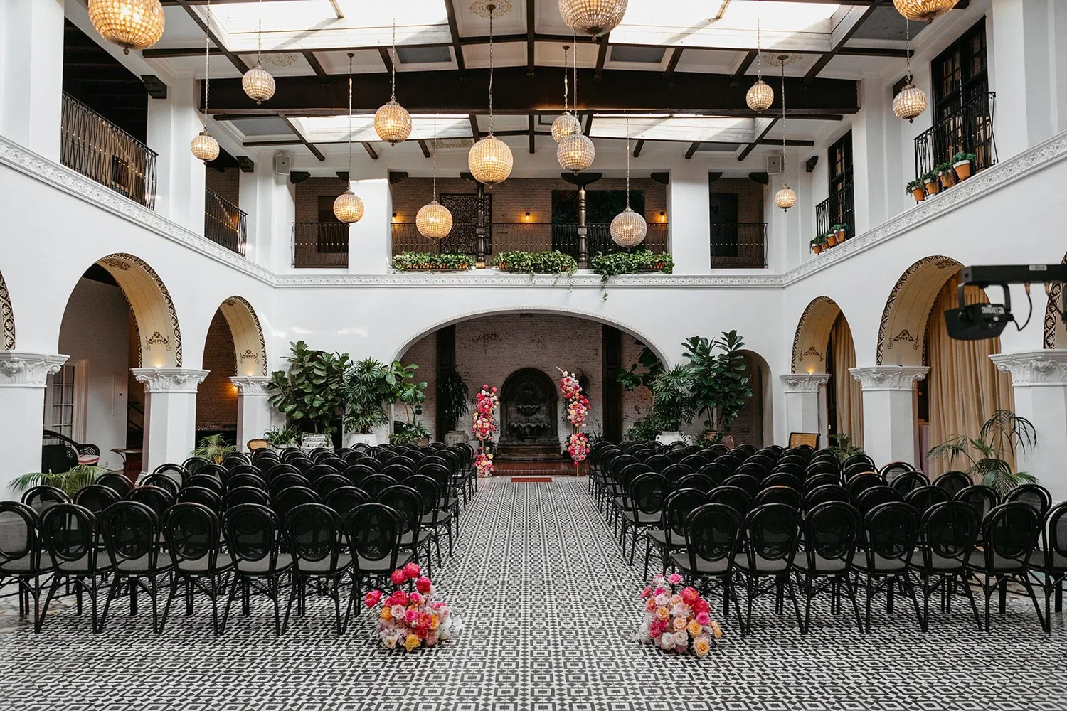Elegant indoor wedding venue with black chairs arranged in rows, floral arrangements on the aisle, decorative plants, and hanging lanterns in a spacious, white-walled hall with a balcony overlooking the main area.