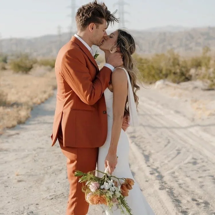 A couple in wedding attire sharing a kiss outdoors, with the man in a rust-colored suit and the woman in a white dress holding a bouquet of flowers, in a desert landscape.