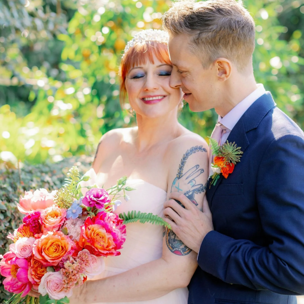 A bride and groom are smiling and leaning their faces together outdoors on a sunny day. The bride has bright red hair, is wearing a white dress, and holding a large colorful bouquet of flowers. The groom is in a dark suit with a boutonniere, and is touching the bride's arm, which has visible tattoos.