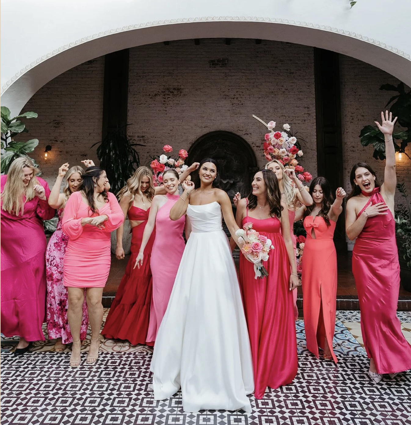 A group of women celebrating at a wedding, with a bride in a white wedding dress and the bridesmaids in various shades of pink and red dresses, holding bouquets of flowers, in an architecturally decorated indoor venue.