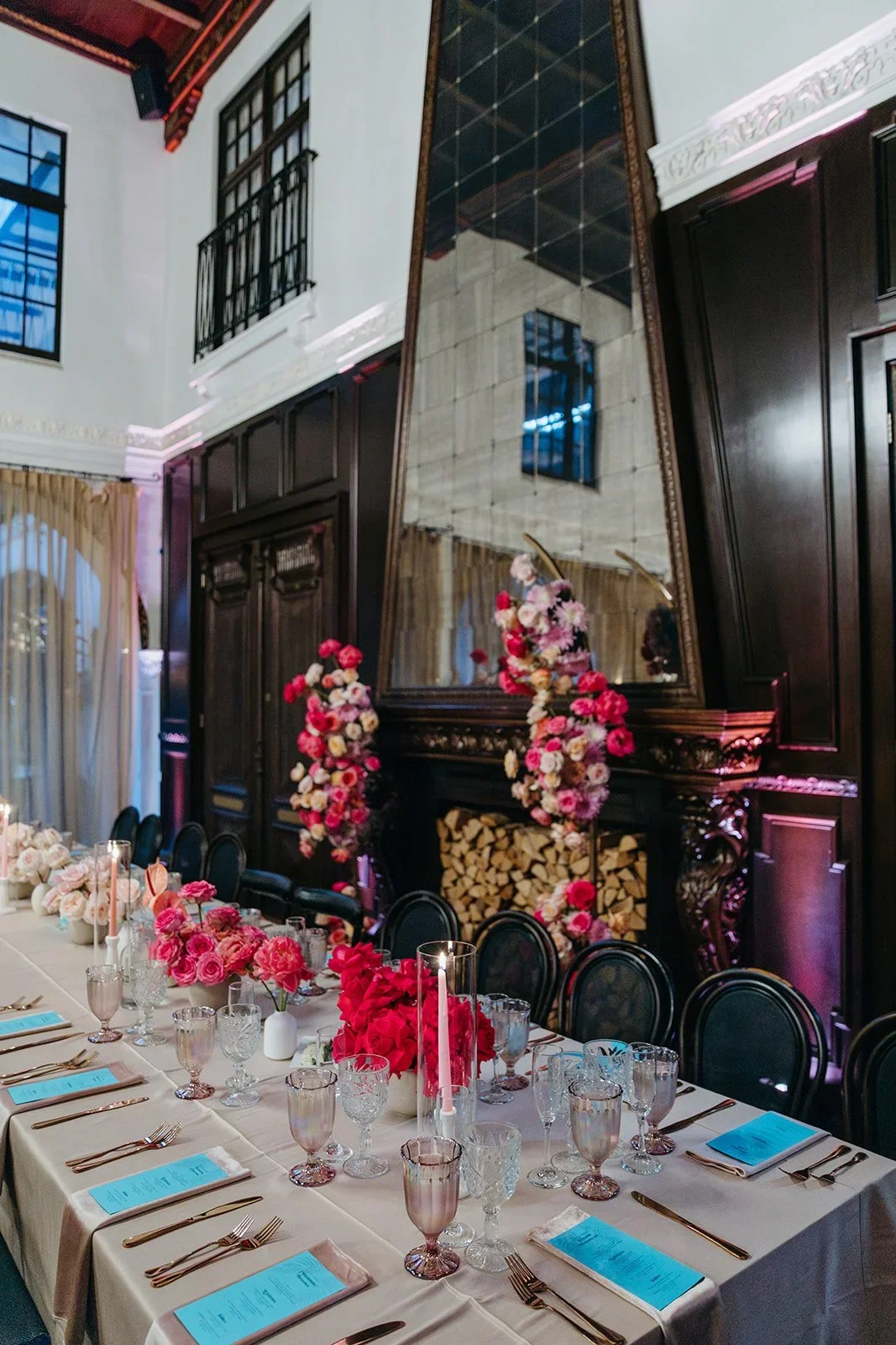 An elegant dining area with a long table set with pink carnations, candles, and glassware. The background features a large mirror above a fireplace with firewood, dark wood paneling, and floral arrangements.
