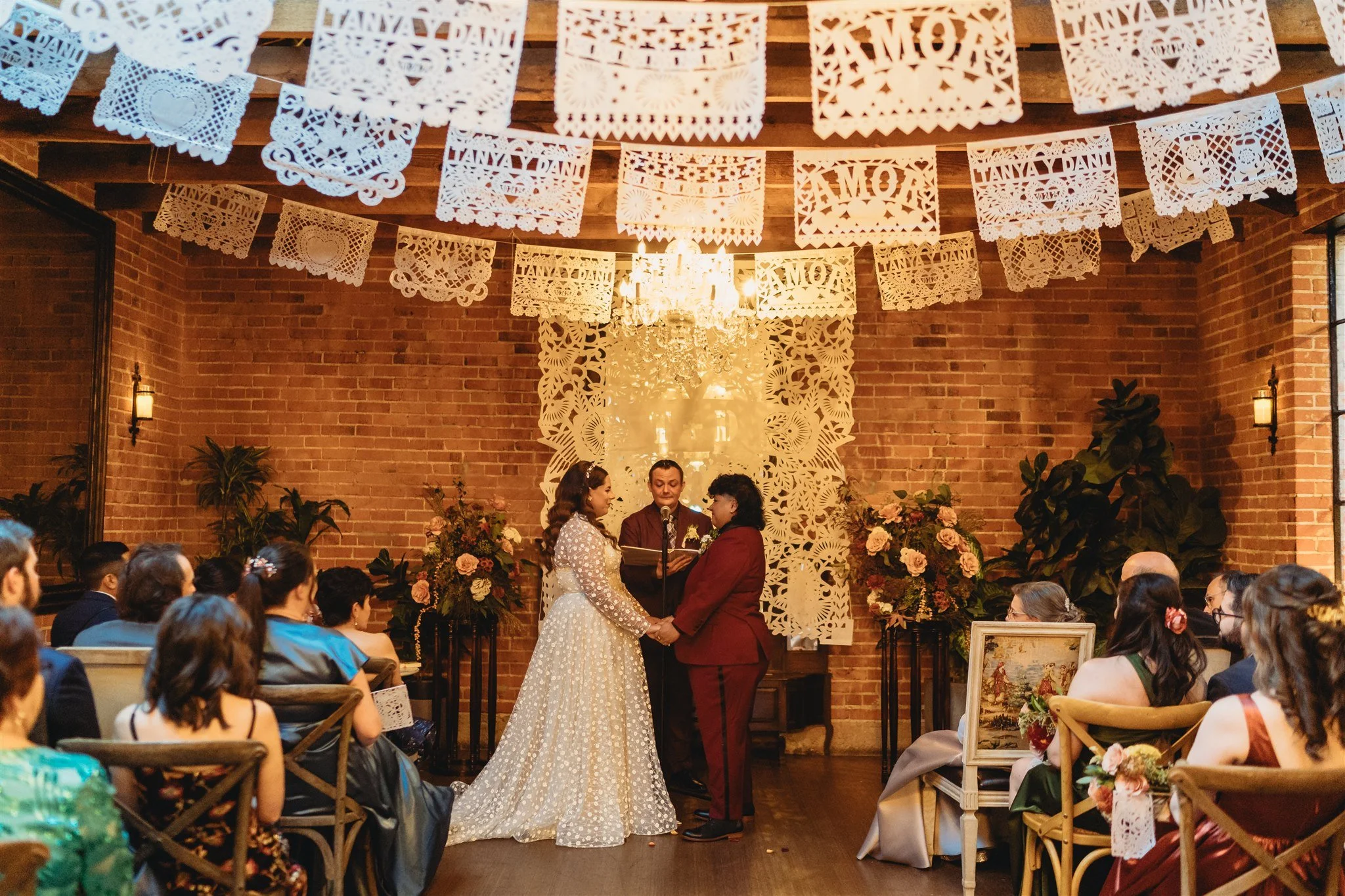 A wedding ceremony with a bride and groom holding hands and facing each other, exchanging vows or rings, in a decorated indoor venue with guests seated and watching. Overhead are decorative paper banners, and a chandelier hangs in the background.