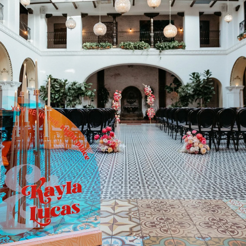 Interior of a wedding or event venue with chairs arranged along a patterned tile aisle, decorated with pink and white flowers, and a floral arch at the front. There are hanging pendant lights, potted plants, and a balcony with more flower arrangements overlooking the space. A sign with the name "Kayla Lucas" is partially visible in the foreground.