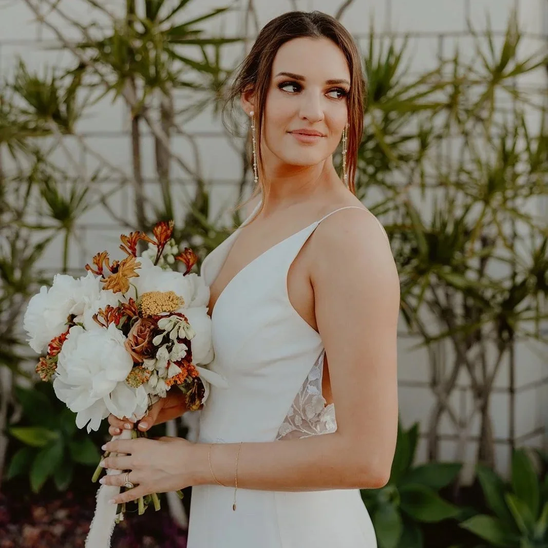 A woman in a white wedding dress holding a bouquet of flowers, standing outdoors with green plants in the background.