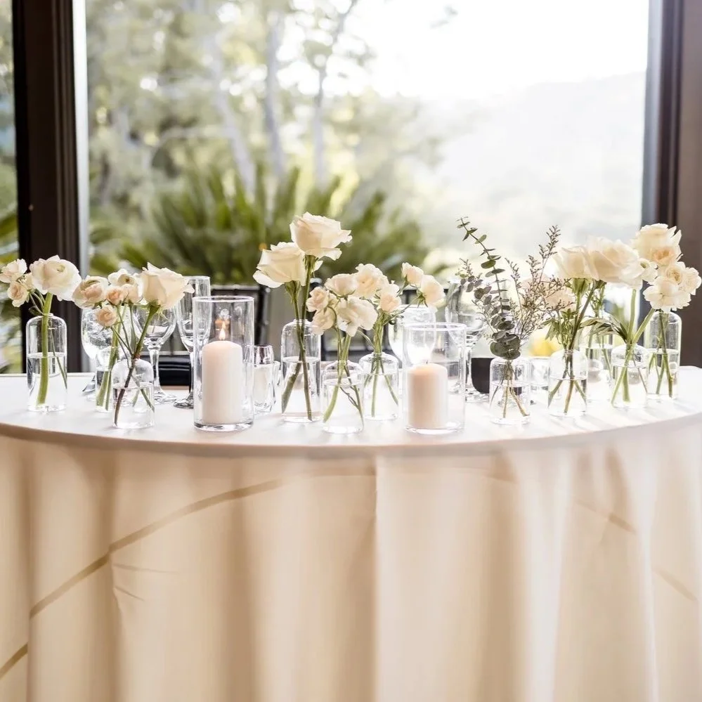 Table decorated with white flowers, glass vases, candles, and greenery, with a window and trees in the background.