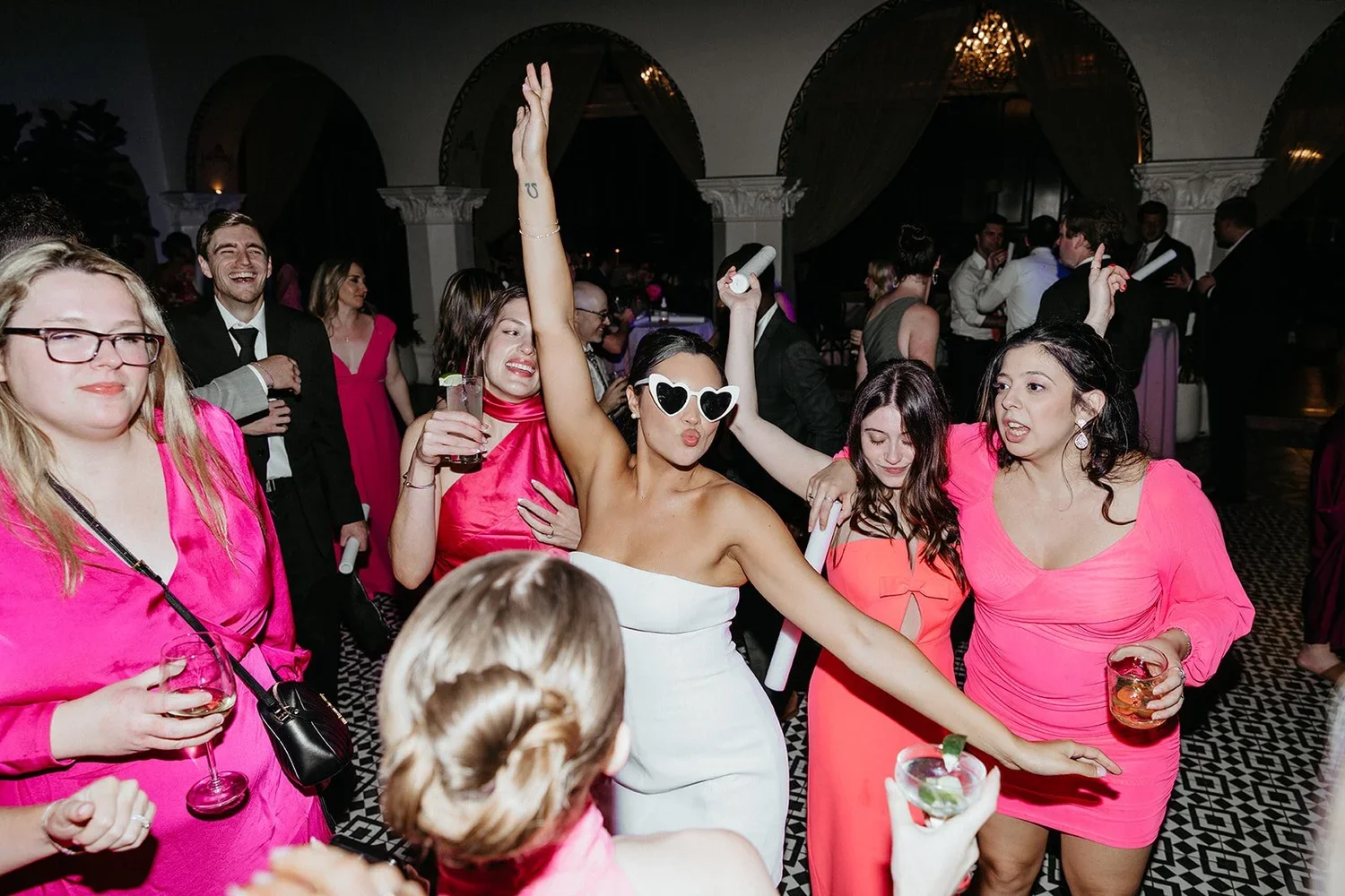 People dancing and having fun at a party, with women wearing pink dresses and a woman in a white strapless dress with sunglasses.