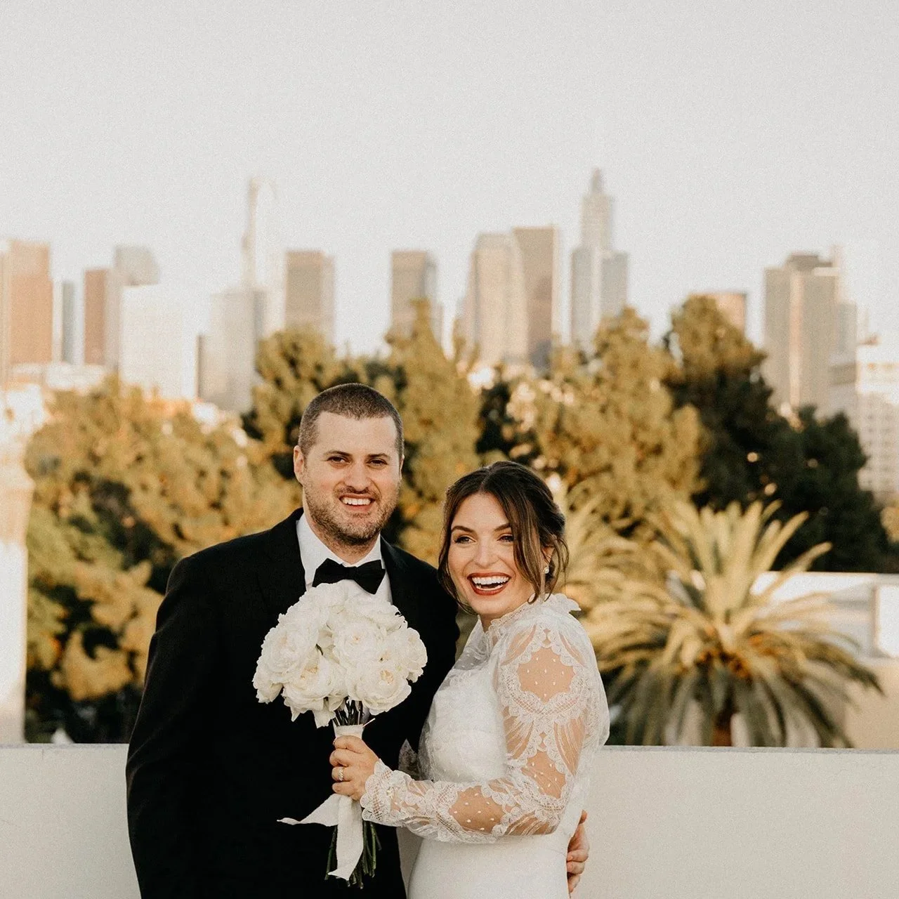 A newlywed couple in wedding attire, smiling and posing outdoors in front of a city skyline with tall buildings and greenery, during sunset.