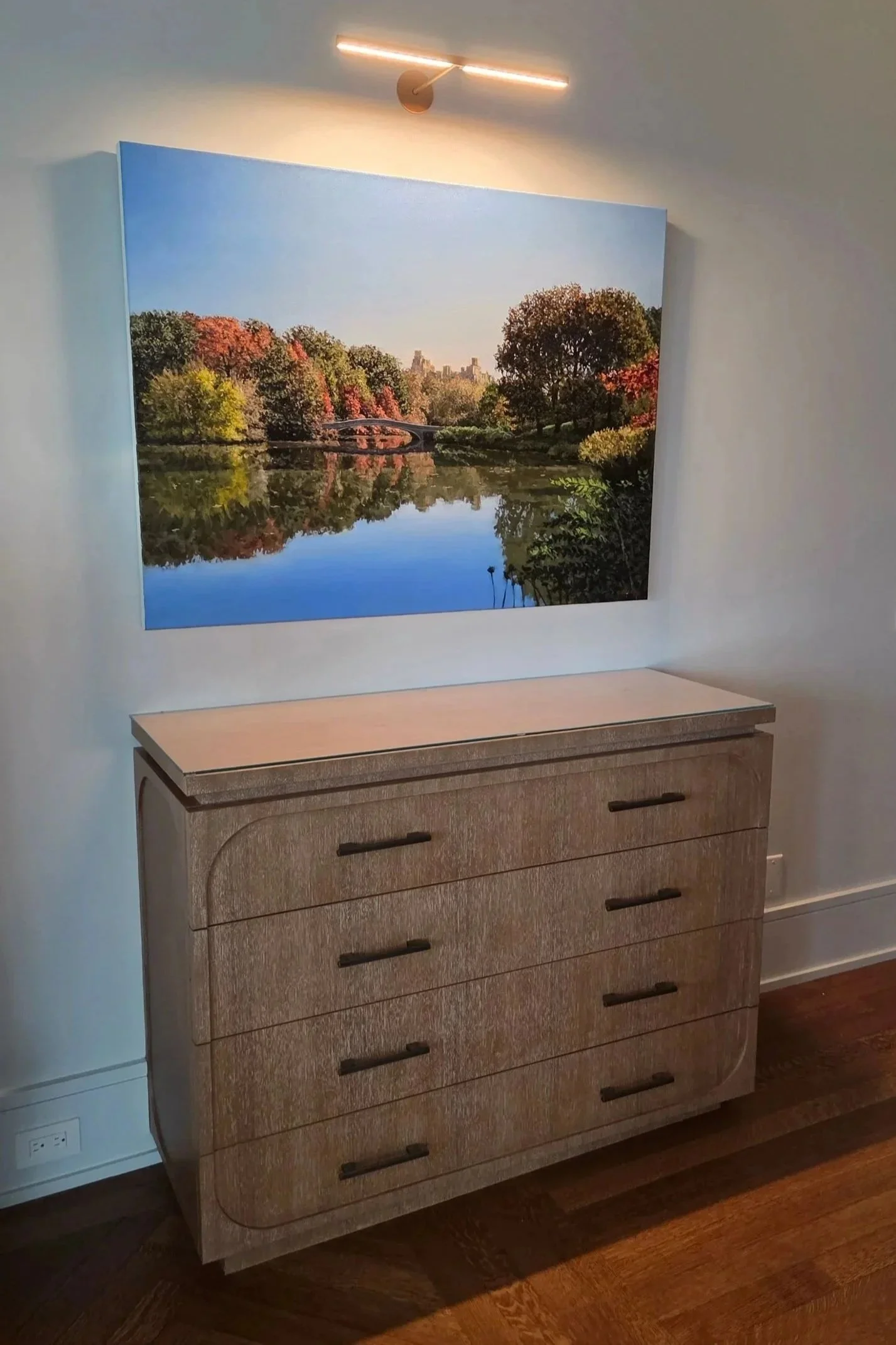 A hallway or living space with a framed landscape photograph of a lake, trees in fall colors, and a bridge, mounted on a wall above a light-colored dresser with black handles.