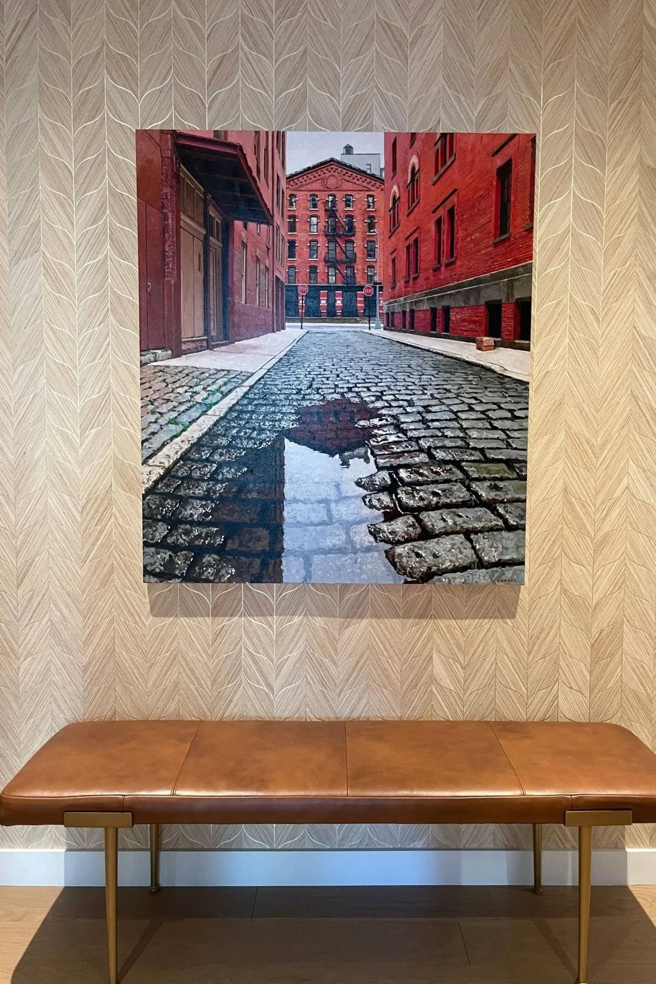 Painting of a cobblestone street with puddles, flanked by red brick buildings under a cloudy sky.