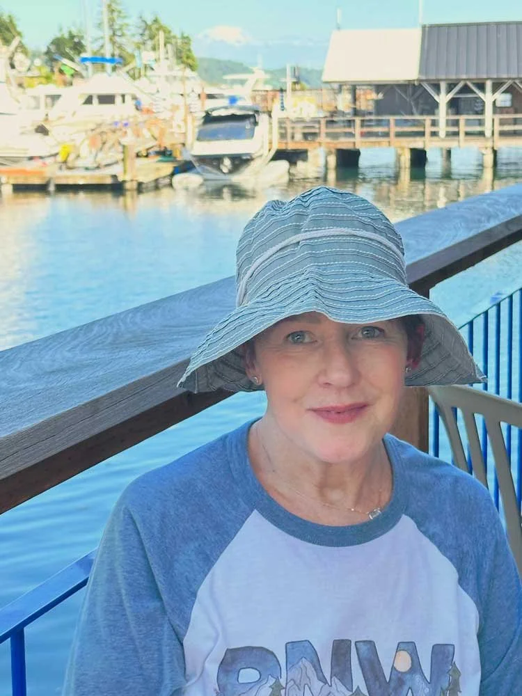 Jennifer Scroggie wearing a wide-brimmed hat and a gray and white baseball-style shirt, sitting near a waterfront in Gig Harbor with boats and a dock in the background.
