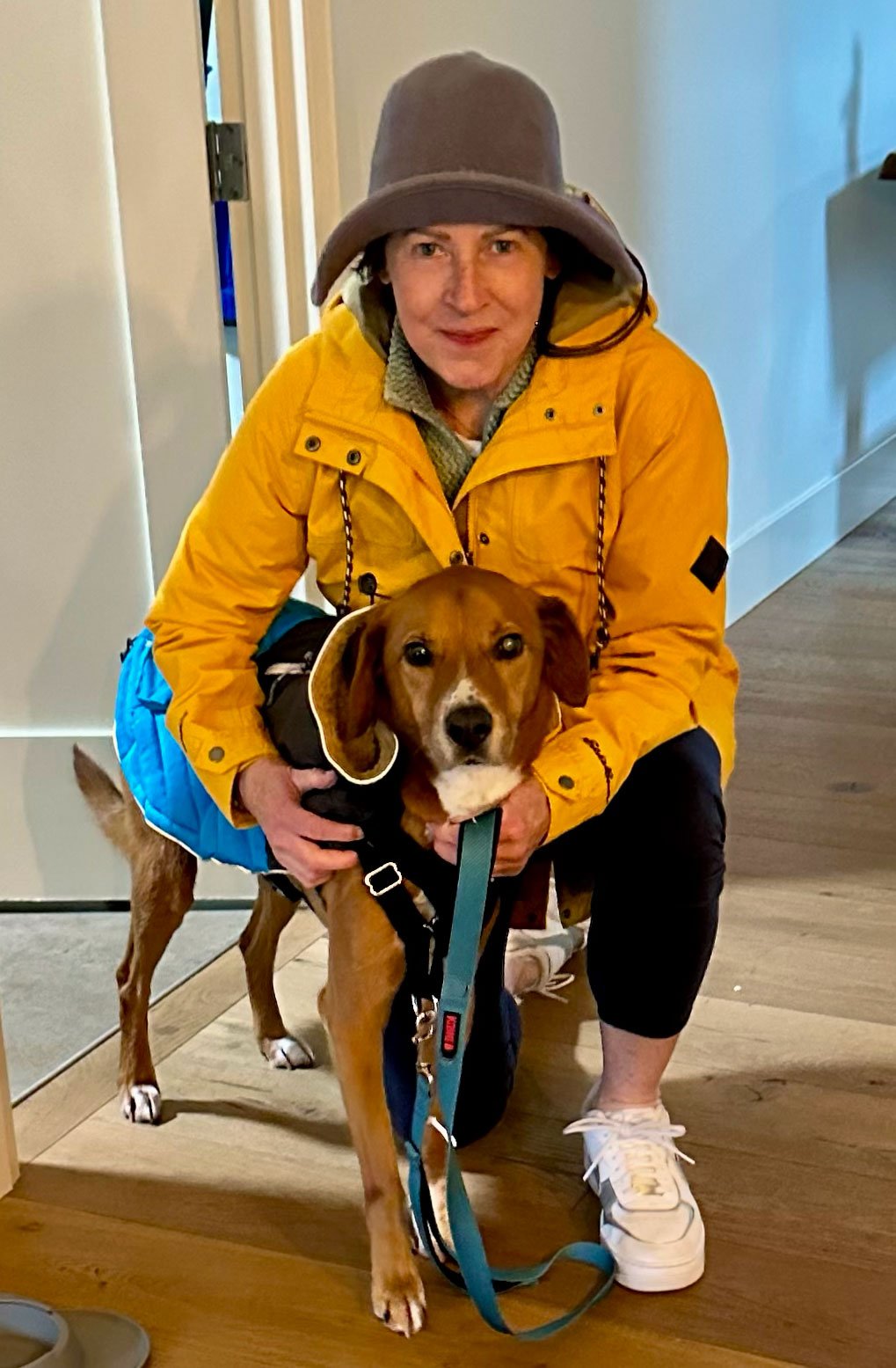 Jennifer Scroggie in a yellow jacket and gray hat crouching beside her dog, Henry.