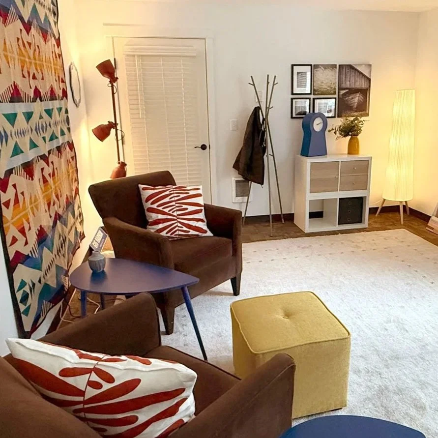 Psychotherapy office for Jennifer Scoggie with brown armchairs, colorful patterned wall hanging, yellow ottoman, small blue side table, framed art on the wall, a coat rack with jackets, a white cabinet with decorative items, and a tall floor lamp.