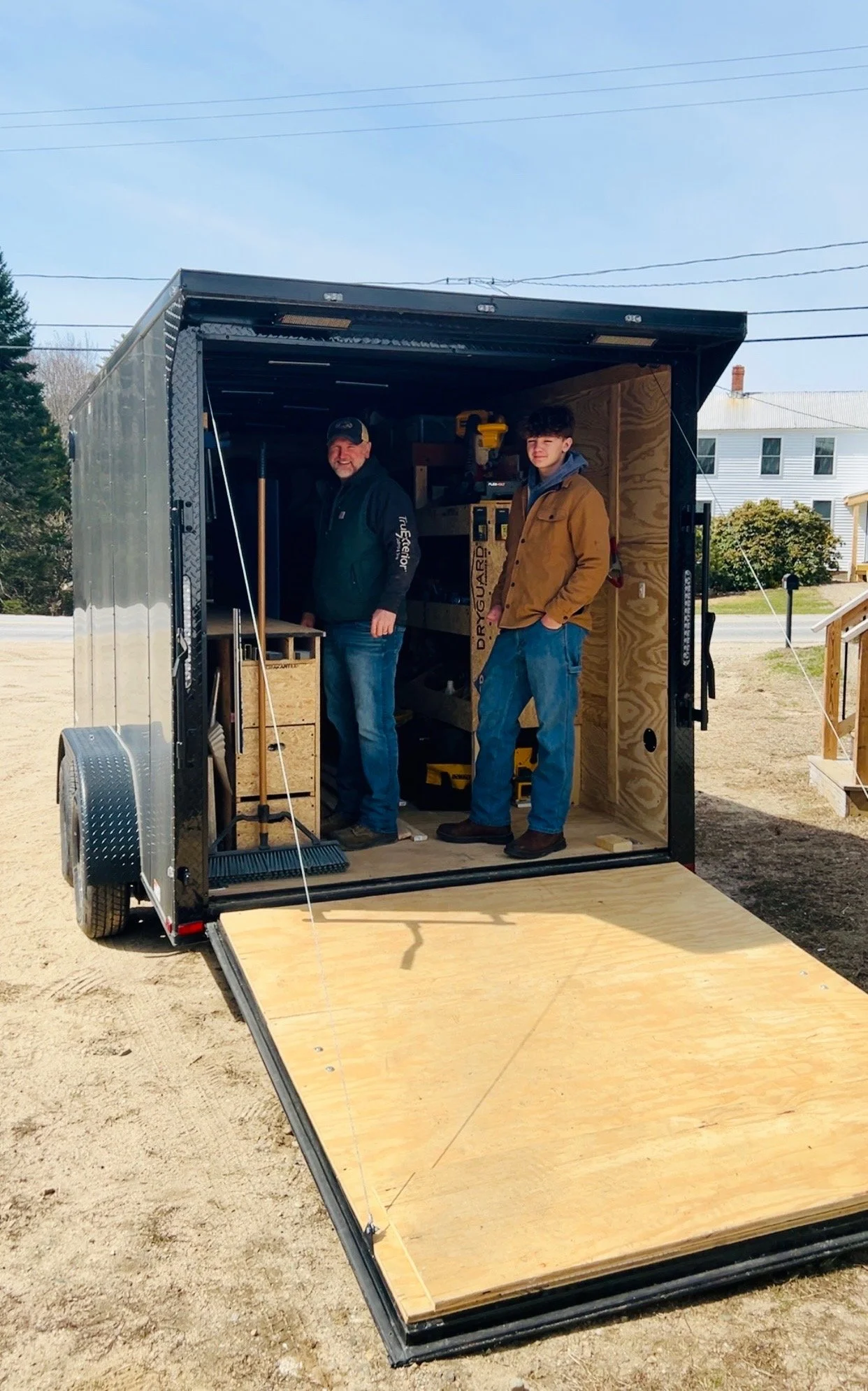 Two men are standing inside a mobile workshop or storage trailer with a wooden floor. One man is smiling and wearing a black jacket and cap, while the other is standing with his hands in his pockets, wearing a brown jacket. The trailer is parked outdoors on a dirt surface, with trees and houses in the background.