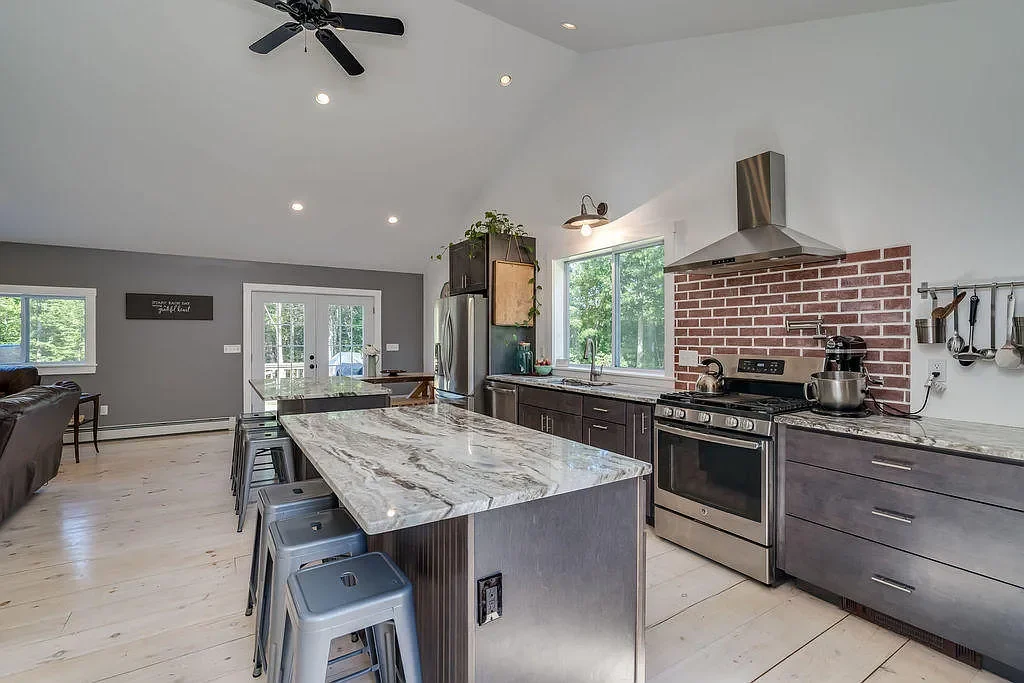 Modern kitchen with gray cabinets, stainless steel appliances, a marble island, and a brick backsplash over the stove. Light wood flooring and large windows with a view of green trees.