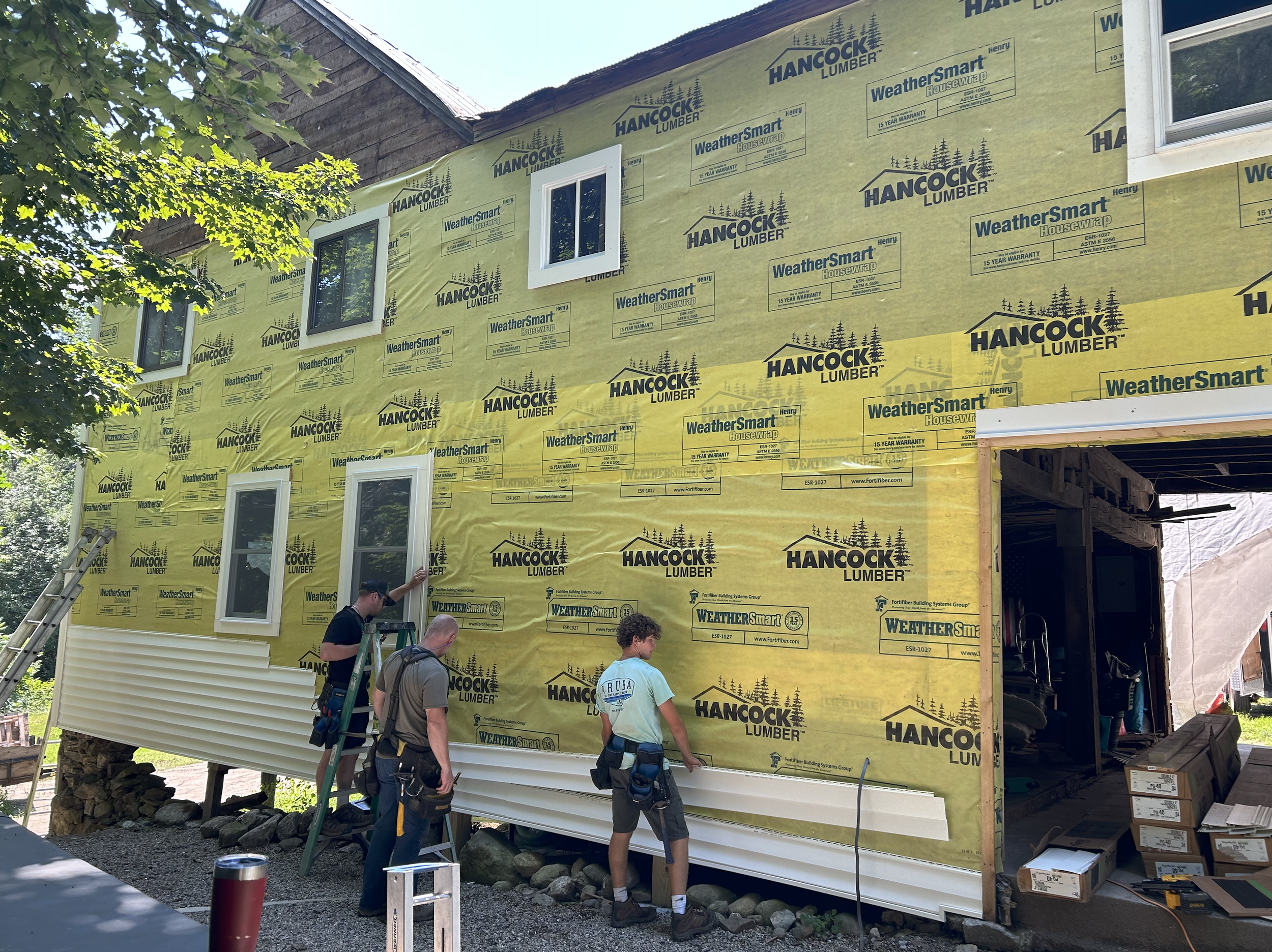 Construction workers working on an unfinished house with yellow weather-resistant siding. One person is on a ladder near the siding, while two others stand nearby holding equipment. The house has windows, and the construction site has boxes and other building materials around.