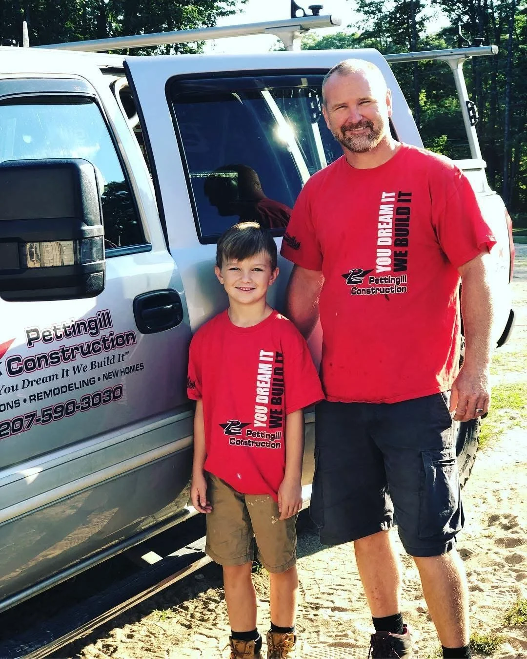 A man and a boy standing outdoors next to a construction company's vehicle, both wearing matching red T-shirts with the company's logo and slogan. The man has a beard and is smiling, while the boy is smiling and has short brown hair. The vehicle advertises Pettingill Construction, with a slogan