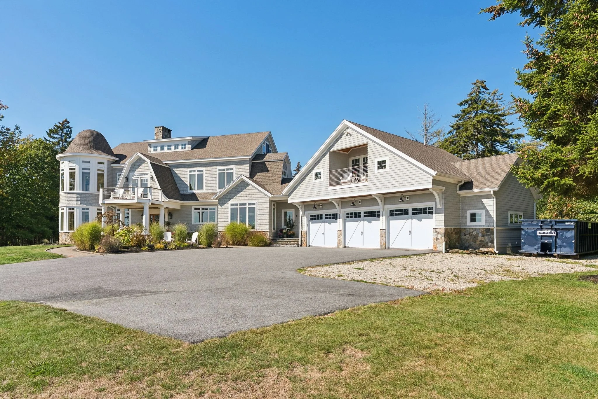 A large, modern house with multiple gabled roofs, white siding, and a wrap-around porch. It has a three-car garage with white doors and a driveway surrounded by a green lawn and shrubs. Tall trees are in the background under a clear blue sky.