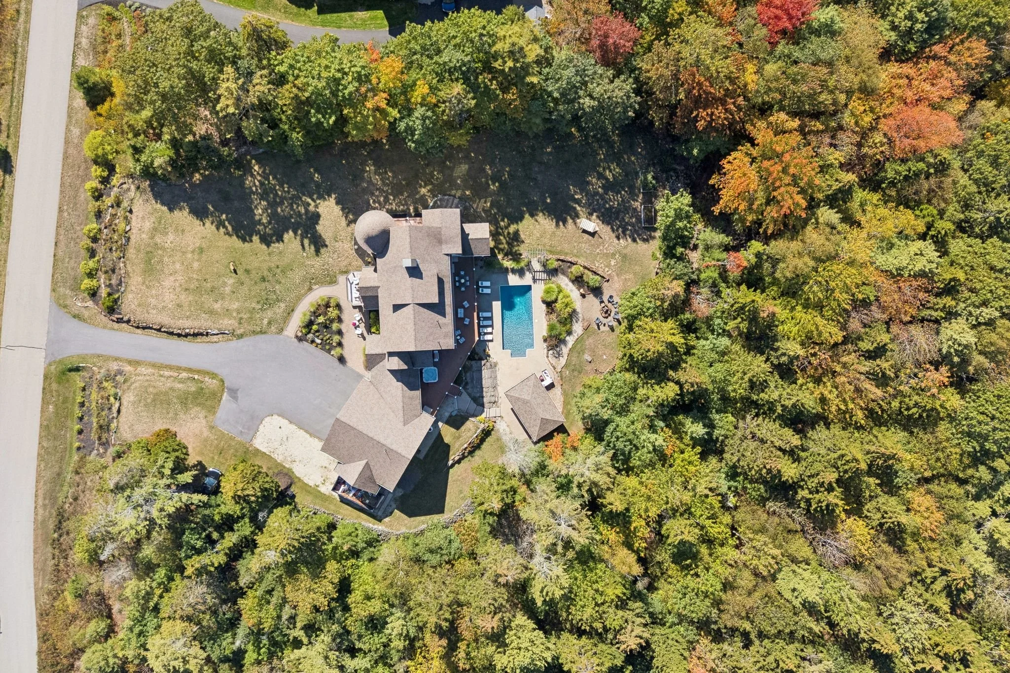 An aerial view of a house with a swimming pool, surrounded by trees with fall foliage, in a neighborhood with a street on the left side.
