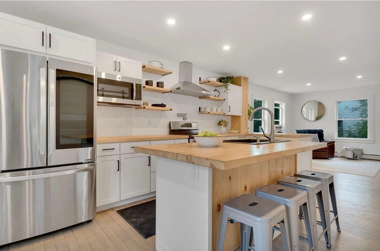 Modern open-concept kitchen with white cabinets, stainless steel appliances, a wooden island with bar stools, and a view of the living room with large windows and a seating area.