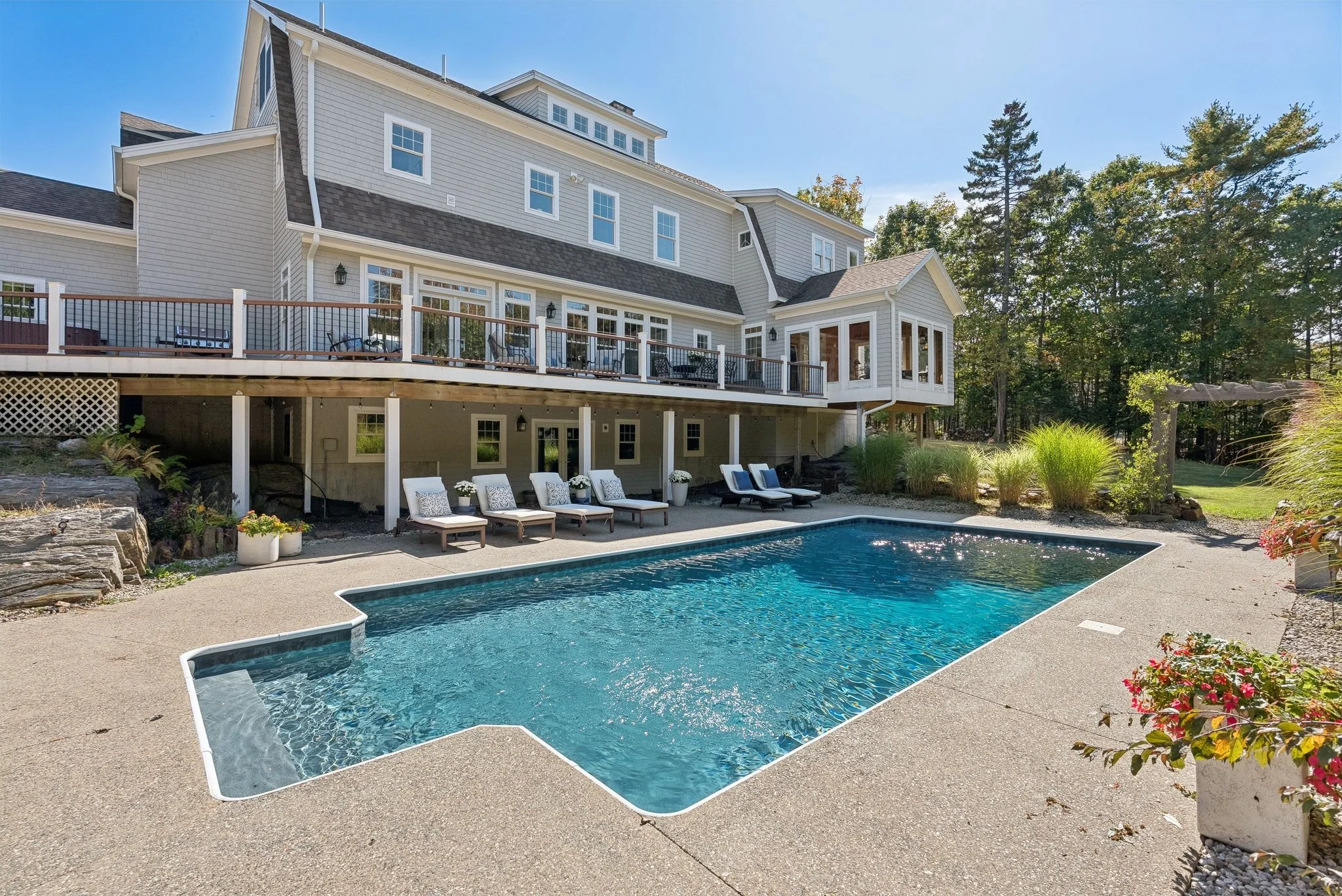 Backyard view of a large house with a swimming pool, patio chairs, and a deck area surrounded by trees and greenery.