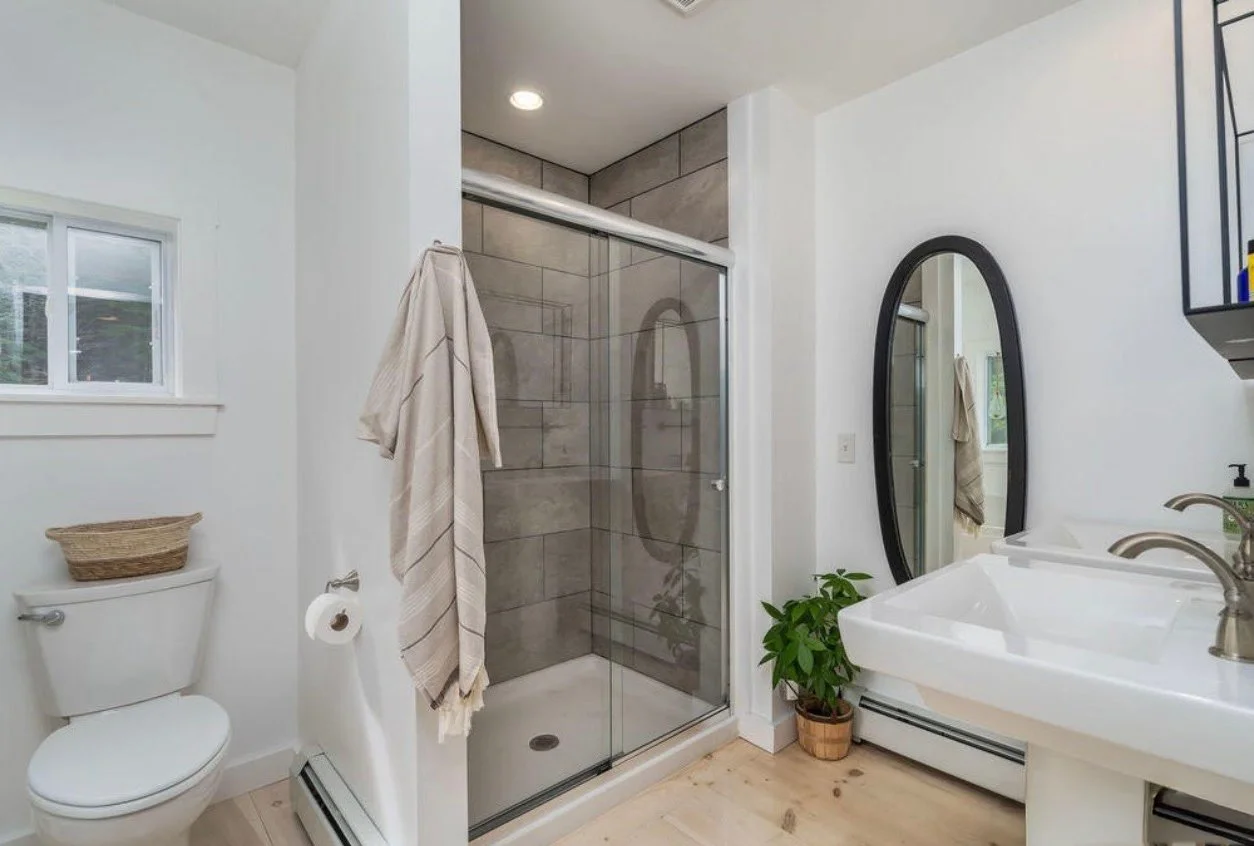 Bathroom with a glass shower enclosure, a white toilet with a basket on top, a potted plant, a white sink, a black oval mirror, a towel hanging, and a window.