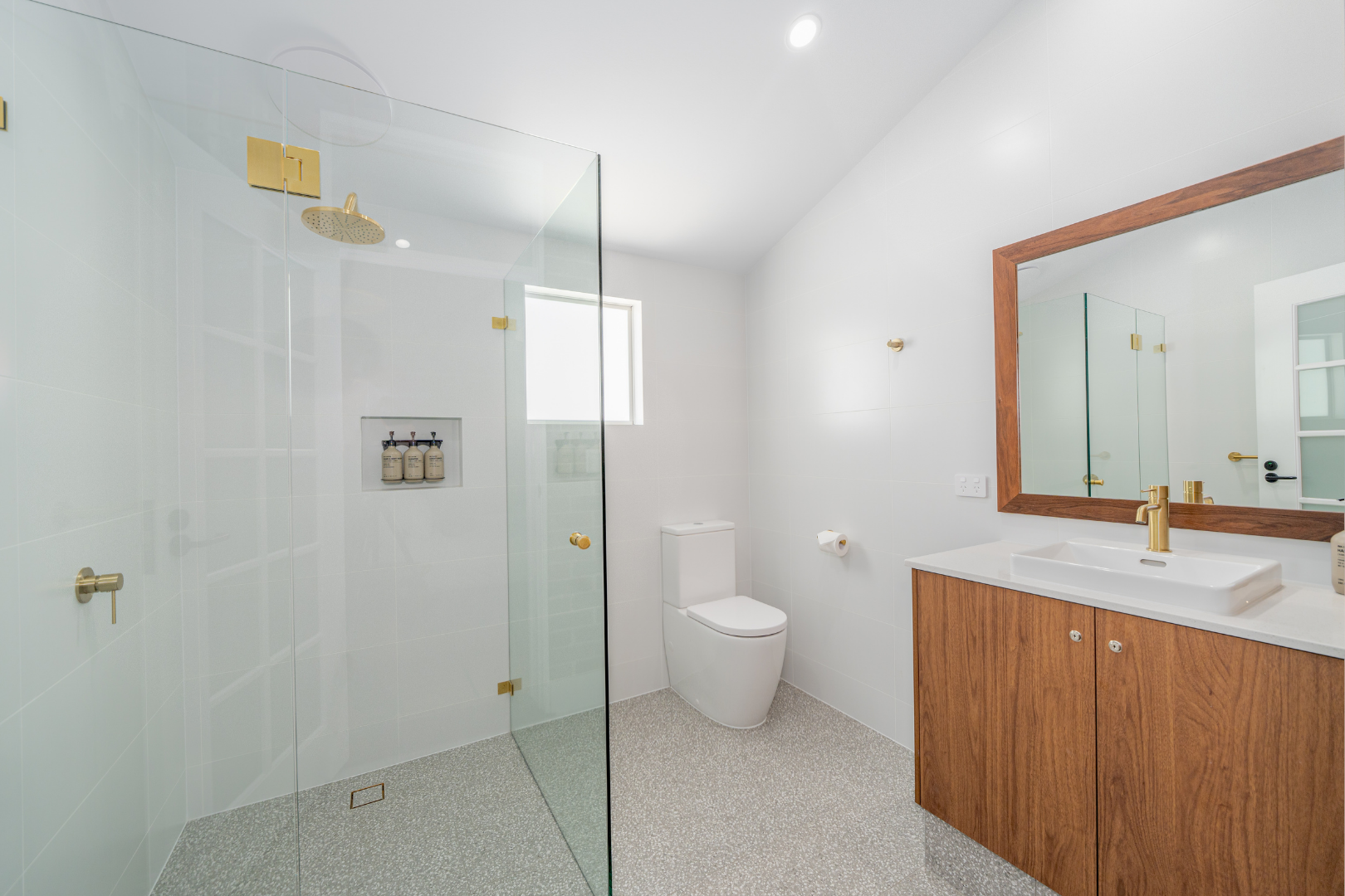 Small modern bathroom with greenish-gray tiled walls, a walk-in shower with a window and blinds, a wooden framed mirror, a black vessel sink on a marble countertop, and a white toilet.