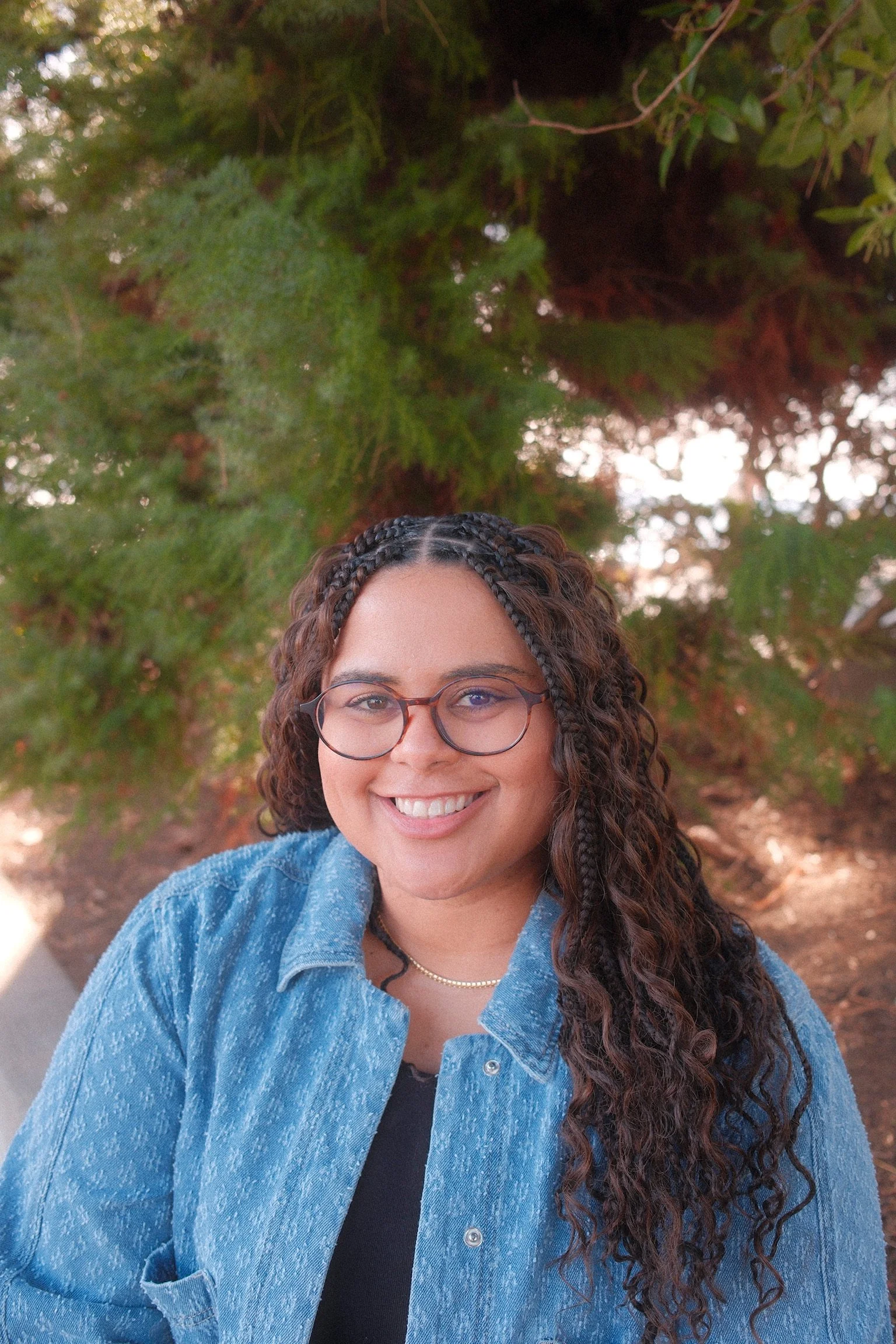 A young woman with curly hair and glasses smiles outdoors, wearing a denim jacket and a black top, with green trees in the background.