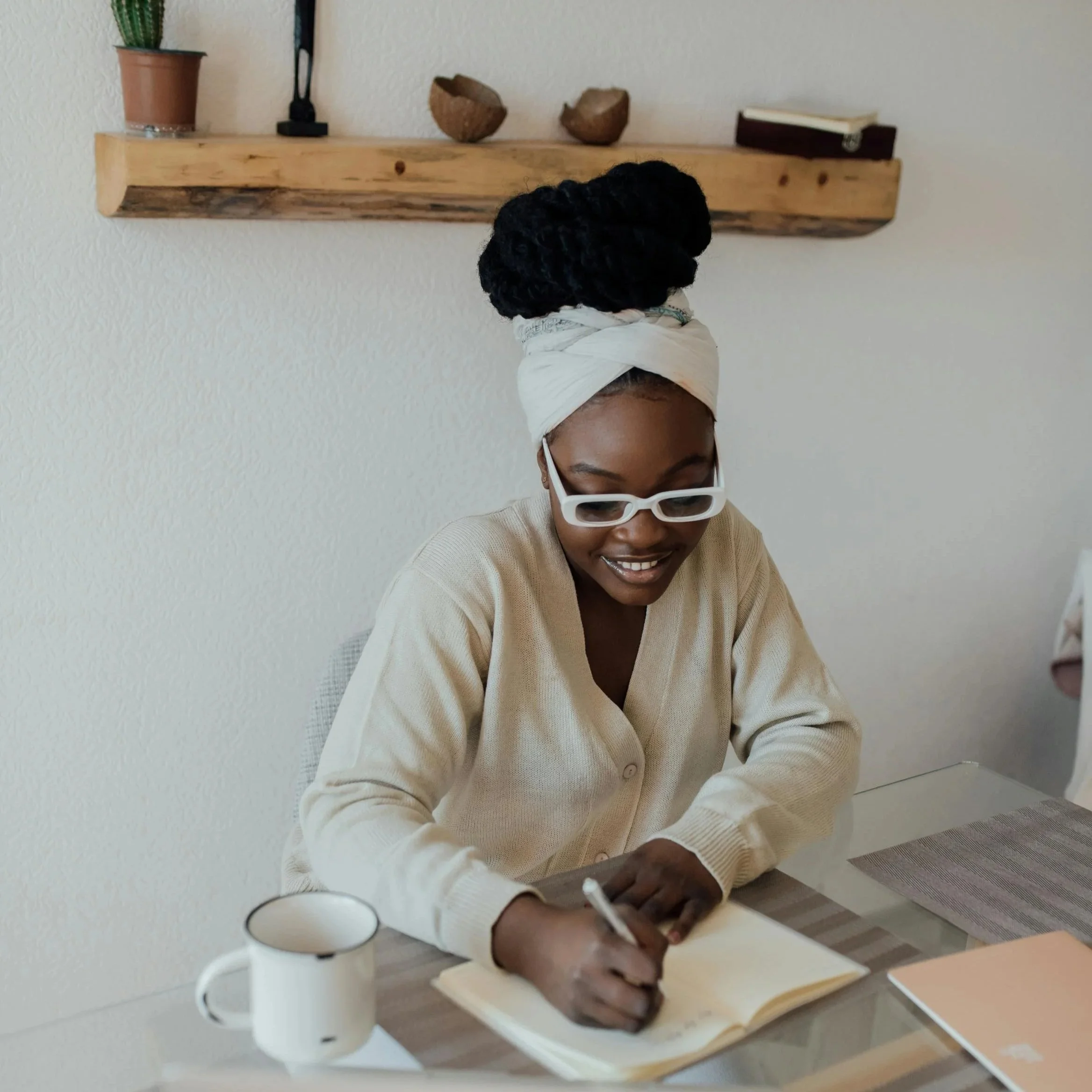 Woman with dark skin, wearing white sunglasses, a beige sweater, and a white headwrap, sitting at a table writing in a notebook, with a white mug nearby and a wooden shelf with decorative objects behind her. Individual Therapy in WA.