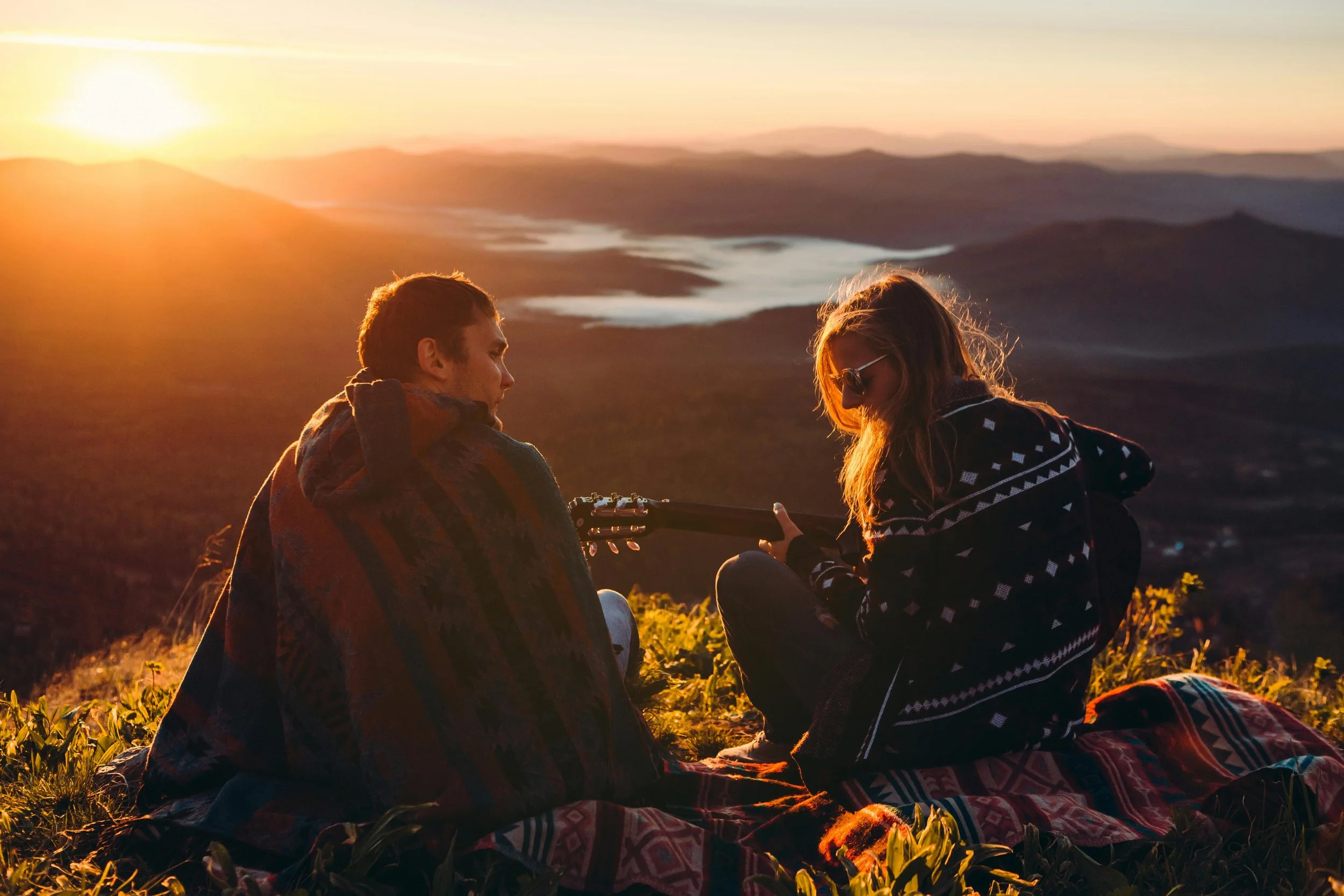 A young man and woman sitting on a blanket on a grassy hill at sunset, with the man holding a guitar and the woman wearing sunglasses, overlooking a scenic landscape of mountains and lakes.