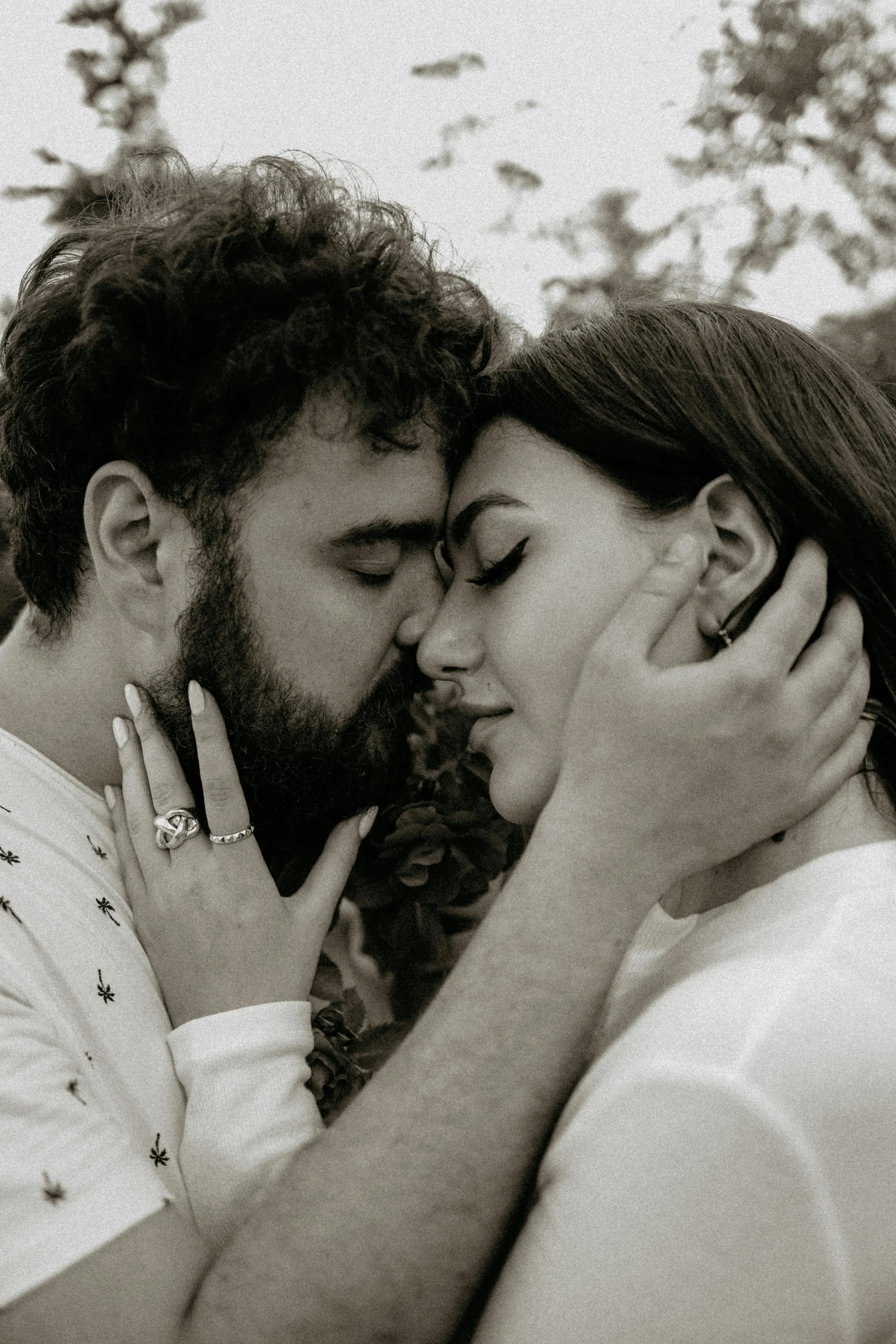 A black and white close-up photo of a couple with eyes closed, gently touching foreheads and faces, outdoors with blurred trees and flowers in the background.