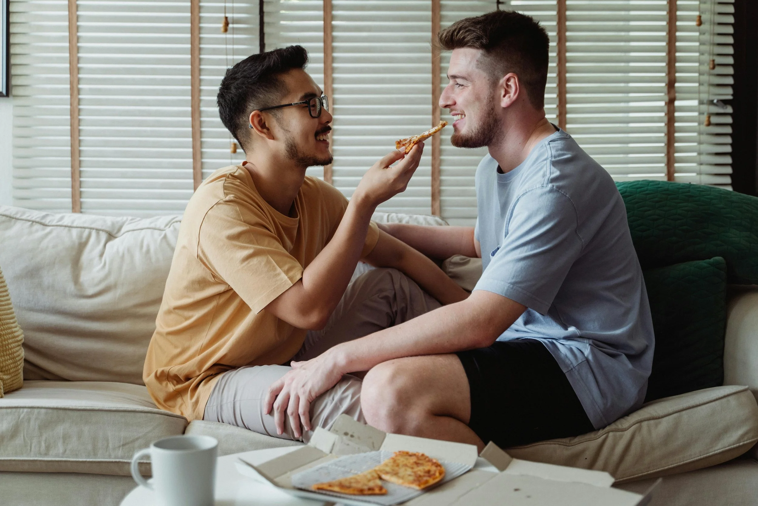 Two men sitting on a couch, one feeding pizza to the other, in a cozy living room with closed blinds.