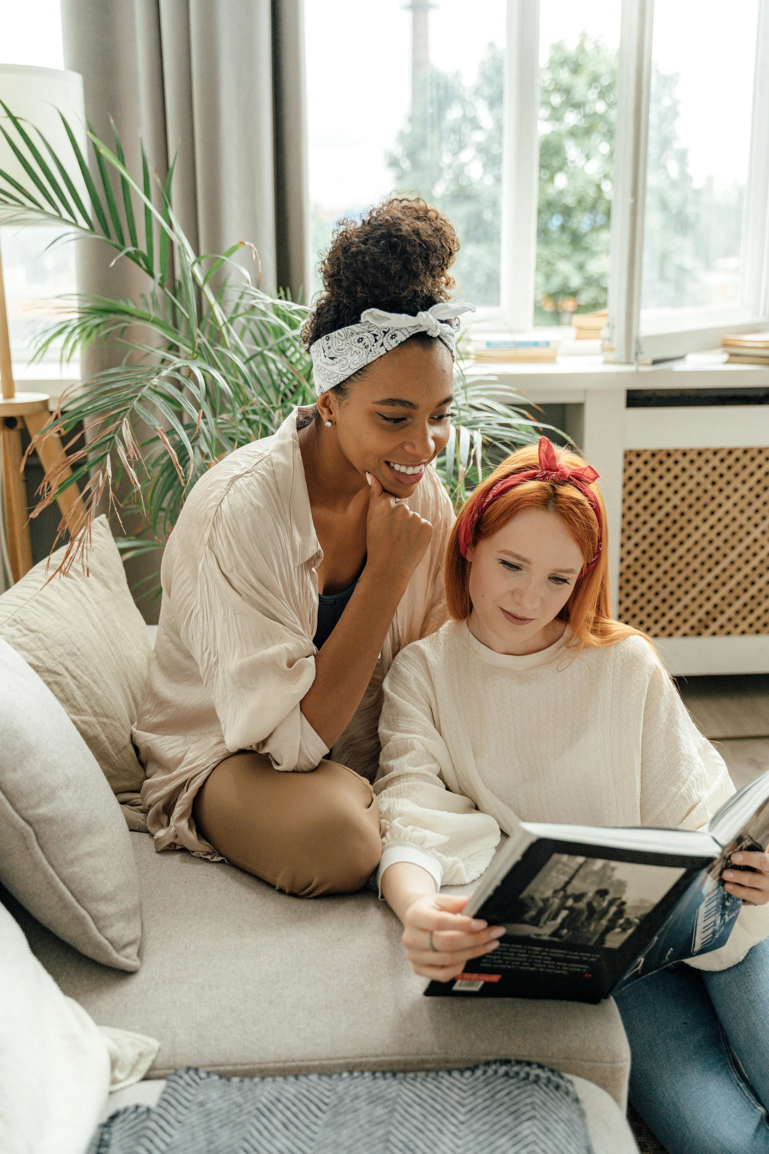Two women sitting on a sofa, one with curly hair and a bandana, the other with red hair and a headband, looking at a book together in a living room. Couples therapy and relationship counseling for Washington State partners