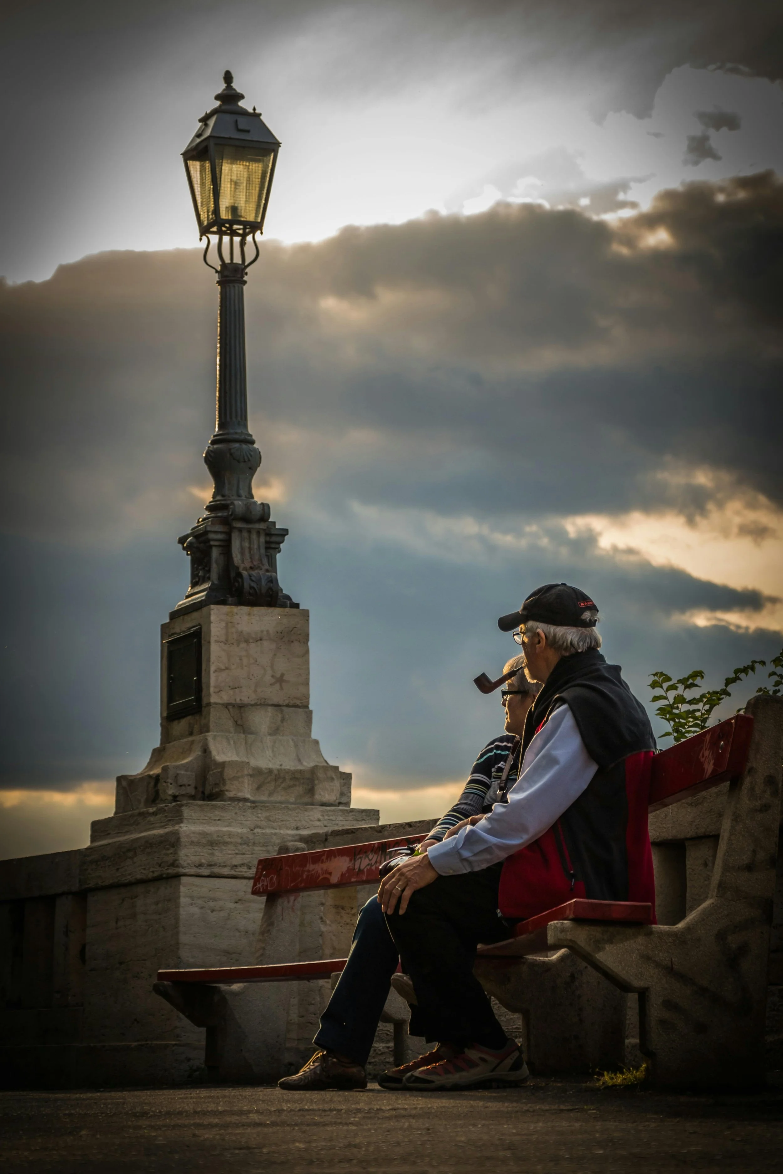 Two elderly people sitting on a bench near a decorative street lamp, with a cloudy sky in the background during sunset. Couples therapy and relationship counseling for Washington State partners