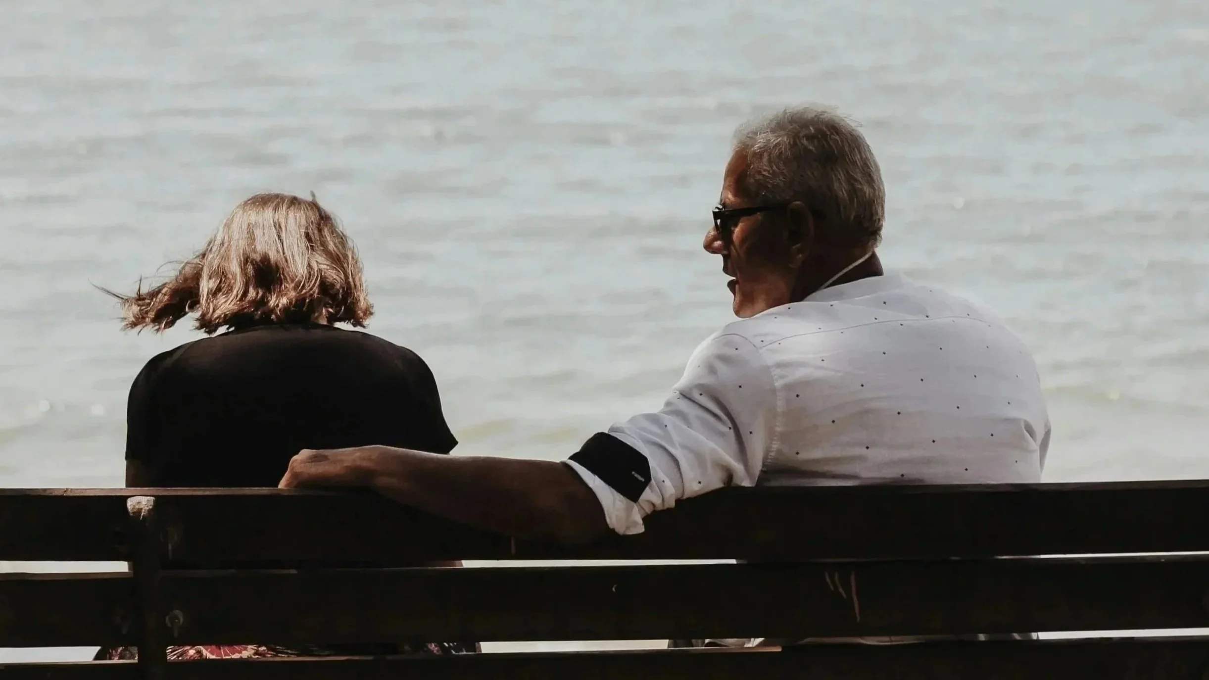 An older man and woman sitting on a bench by the water, engaged in conversation.