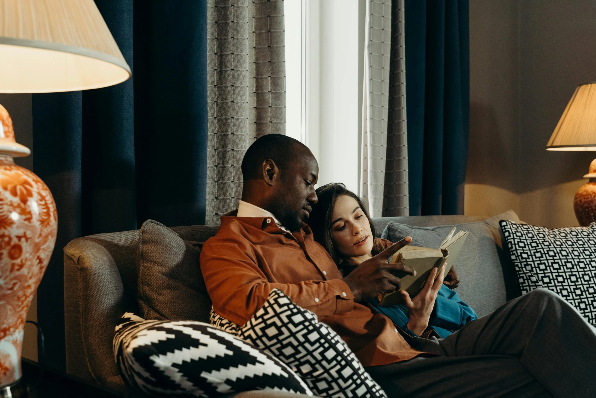 A man and woman sitting on a sofa, looking at a book together in a cozy living room with curtains and lamps. Couples therapy and relationship counseling for Washington State partners