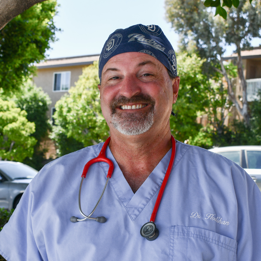A smiling male healthcare professional wearing light blue scrubs, a red stethoscope, and a blue bandana with white logos, standing outdoors with trees and a building in the background.