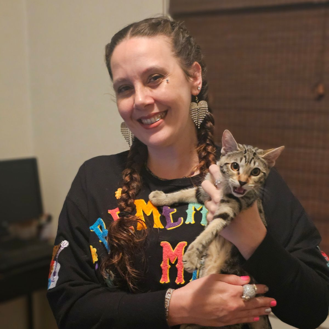 A woman with braided hair, wearing festive earrings and a black shirt with colorful lettering, is smiling and holding a tabby cat with a surprised expression.