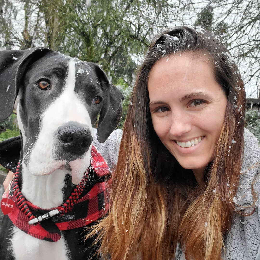 A woman and a black and white dog taking a selfie outdoors in a snowy setting with trees in the background.