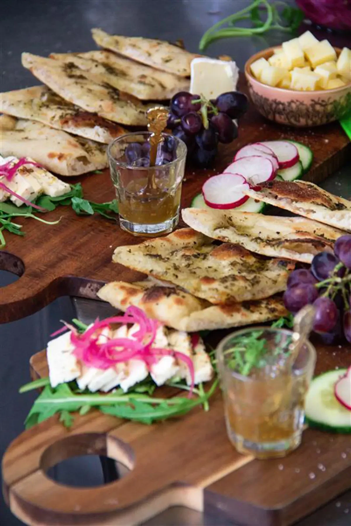 Eine Holzplatte mit Fladenbrot, Radieschen, Gurken, Käse, Trauben, einem Glas Honig und einer Käseplatte mit Frühlingszwiebeln und Honig.