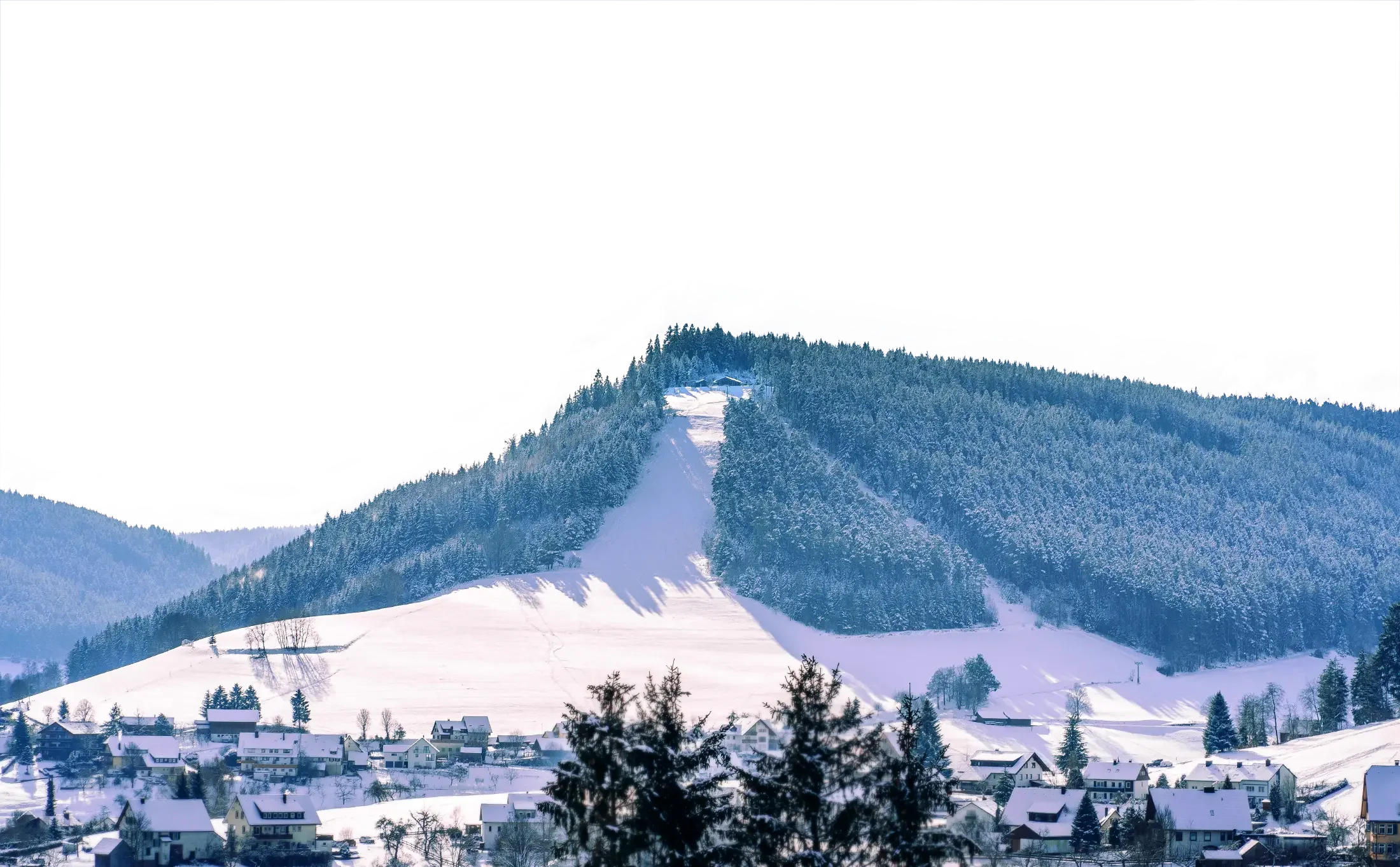 Schneebedeutes Dorf am Fuße eines bewaldeten Hügels mit Skilift und steiler Piste