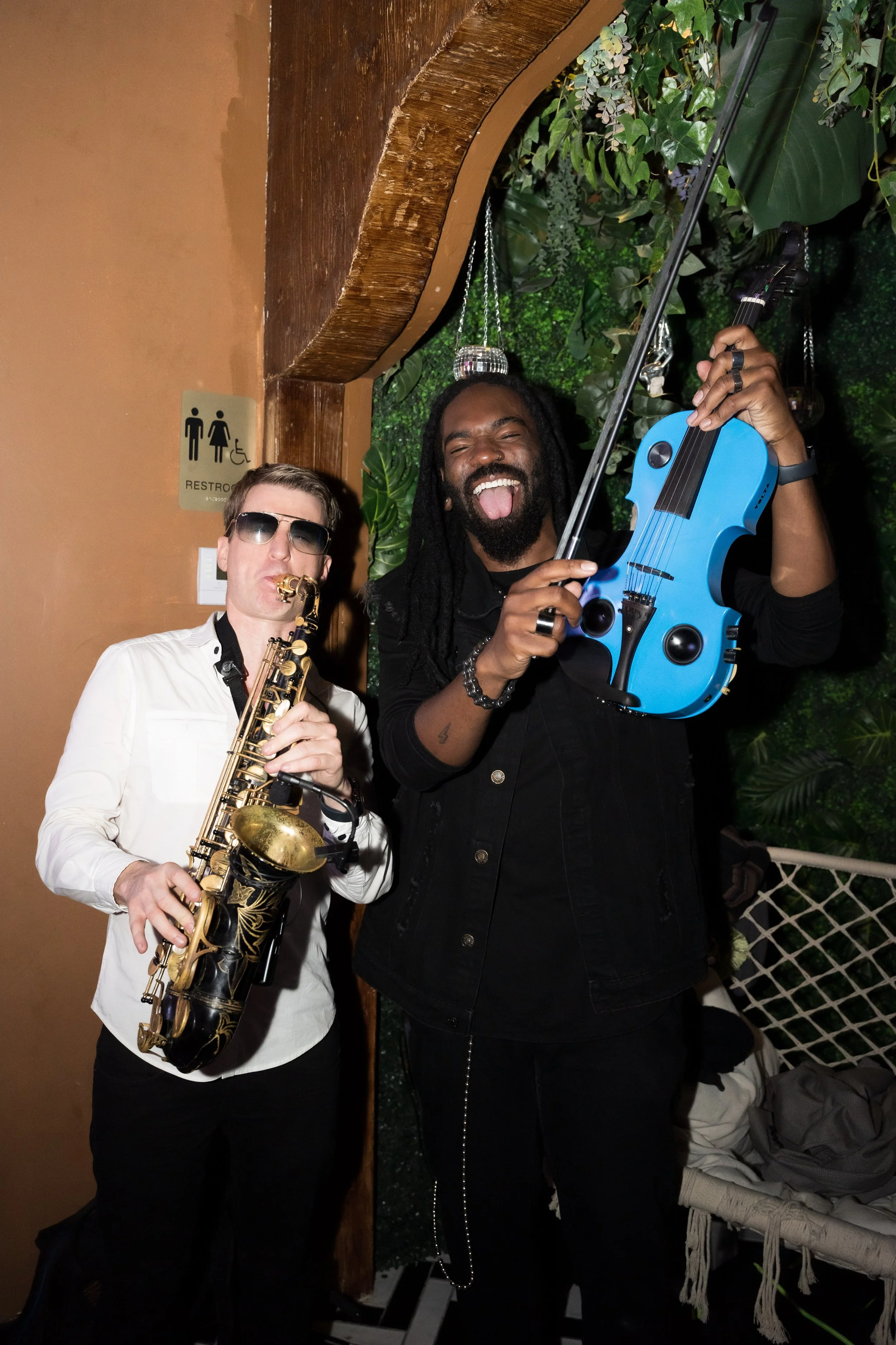 Two musicians posing with their instruments, a man holding a blue electric violin and a woman with sunglasses playing a gold saxophone, smiling and making funny faces in a lively setting with greenery in the background.
