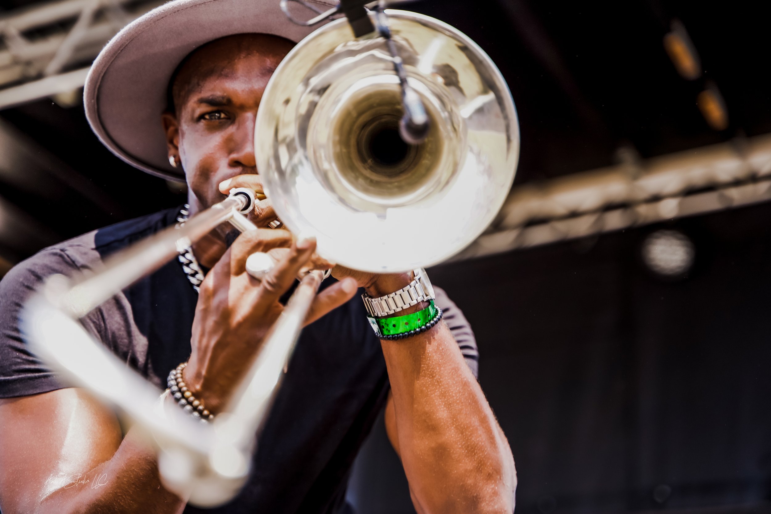 A person wearing a large hat and various bracelets, playing a silver trumpet, taken from a low angle focusing on the instrument's bell.